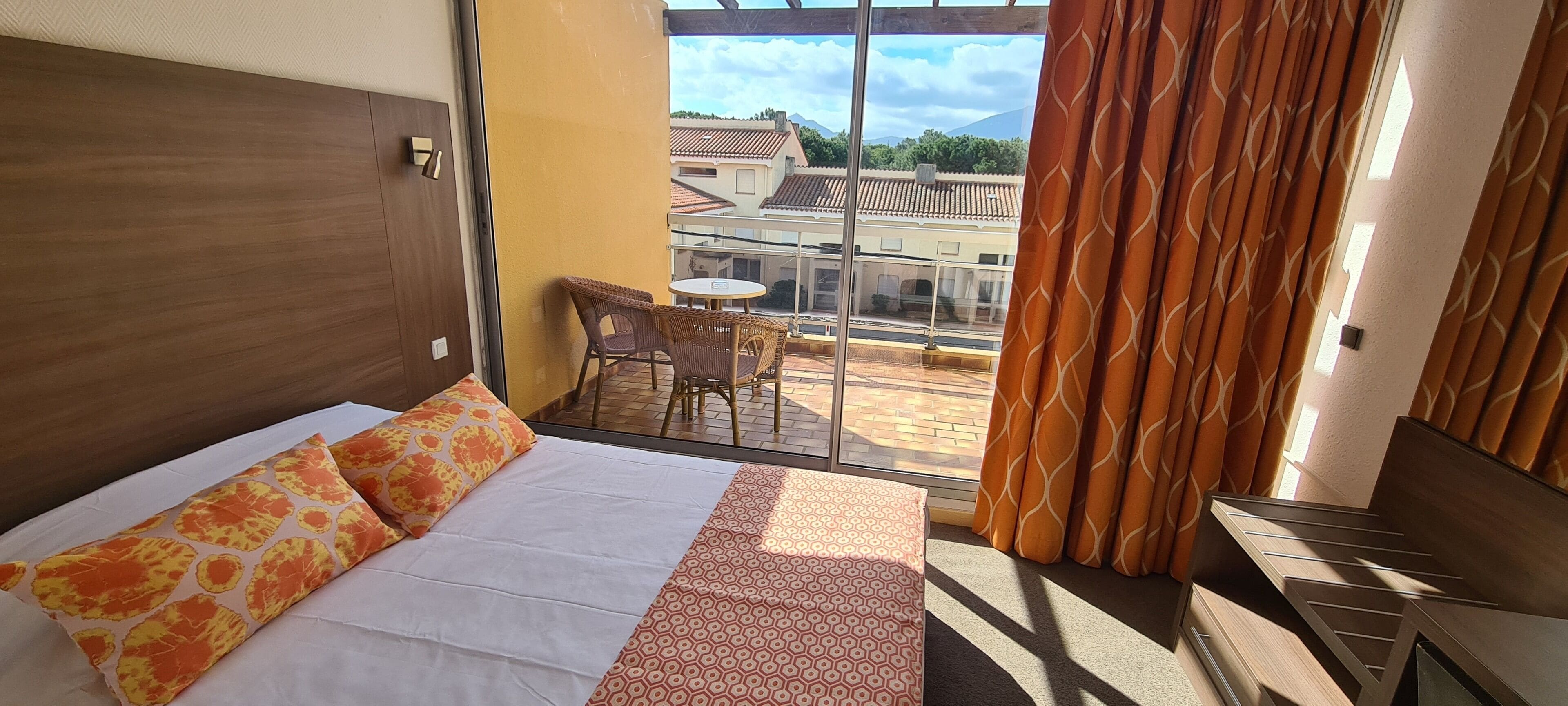 A hotel room with a bed, a small balcony with wicker chairs, and a view of buildings.