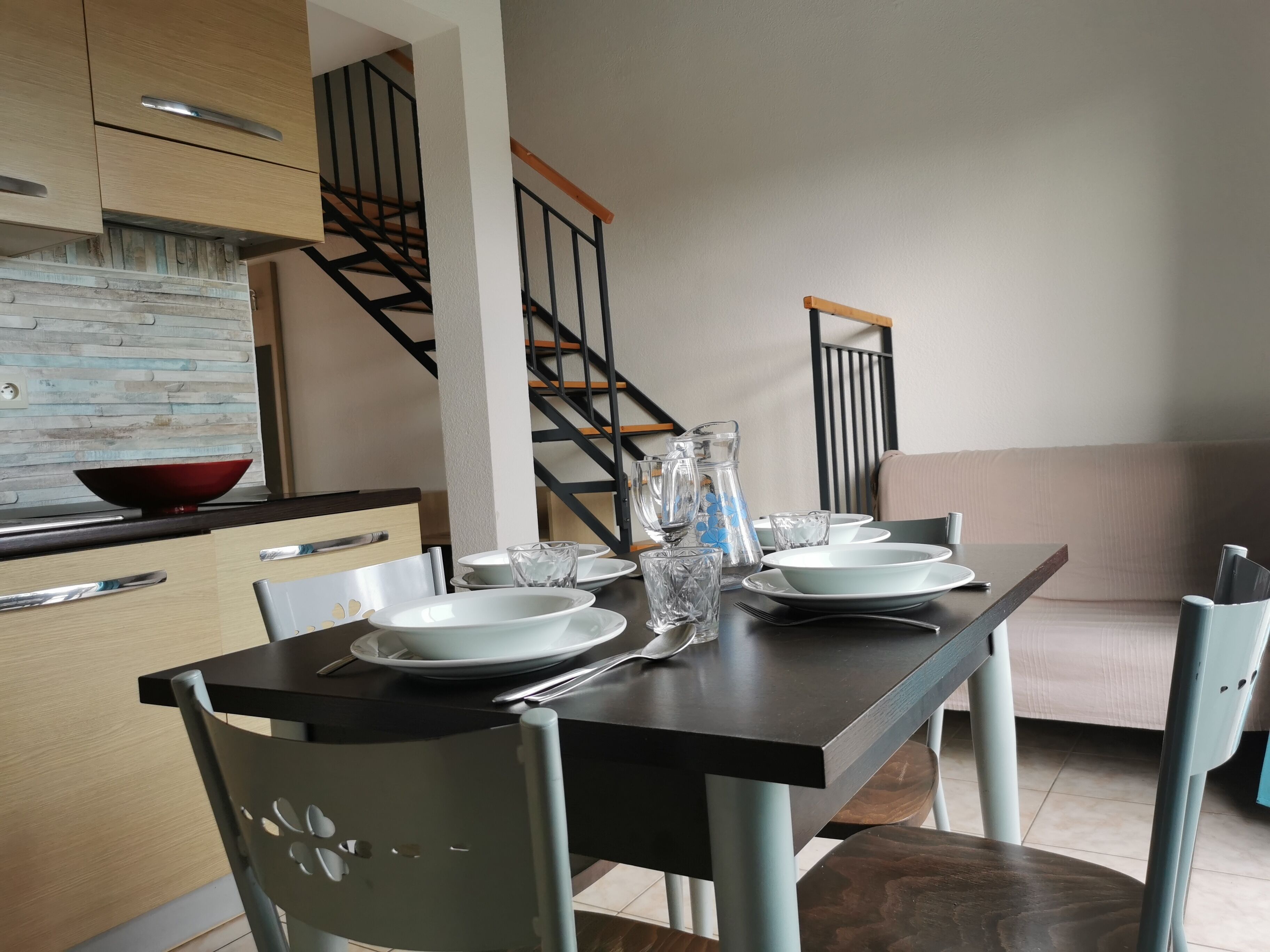 A dining area with a table set for four, a staircase, and a kitchen area with a red bowl on the counter.
