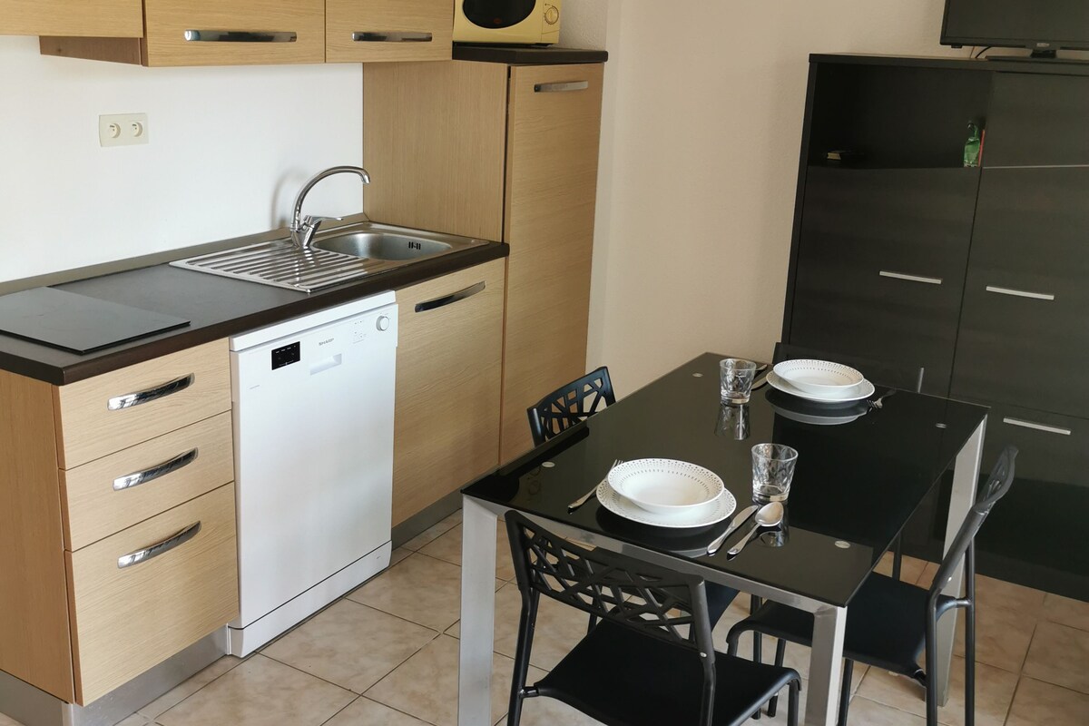 Une cuisine compacte avec des meubles en bois, un lave-vaisselle, une table à manger noire avec des chaises et un téléviseur posé sur un meuble.