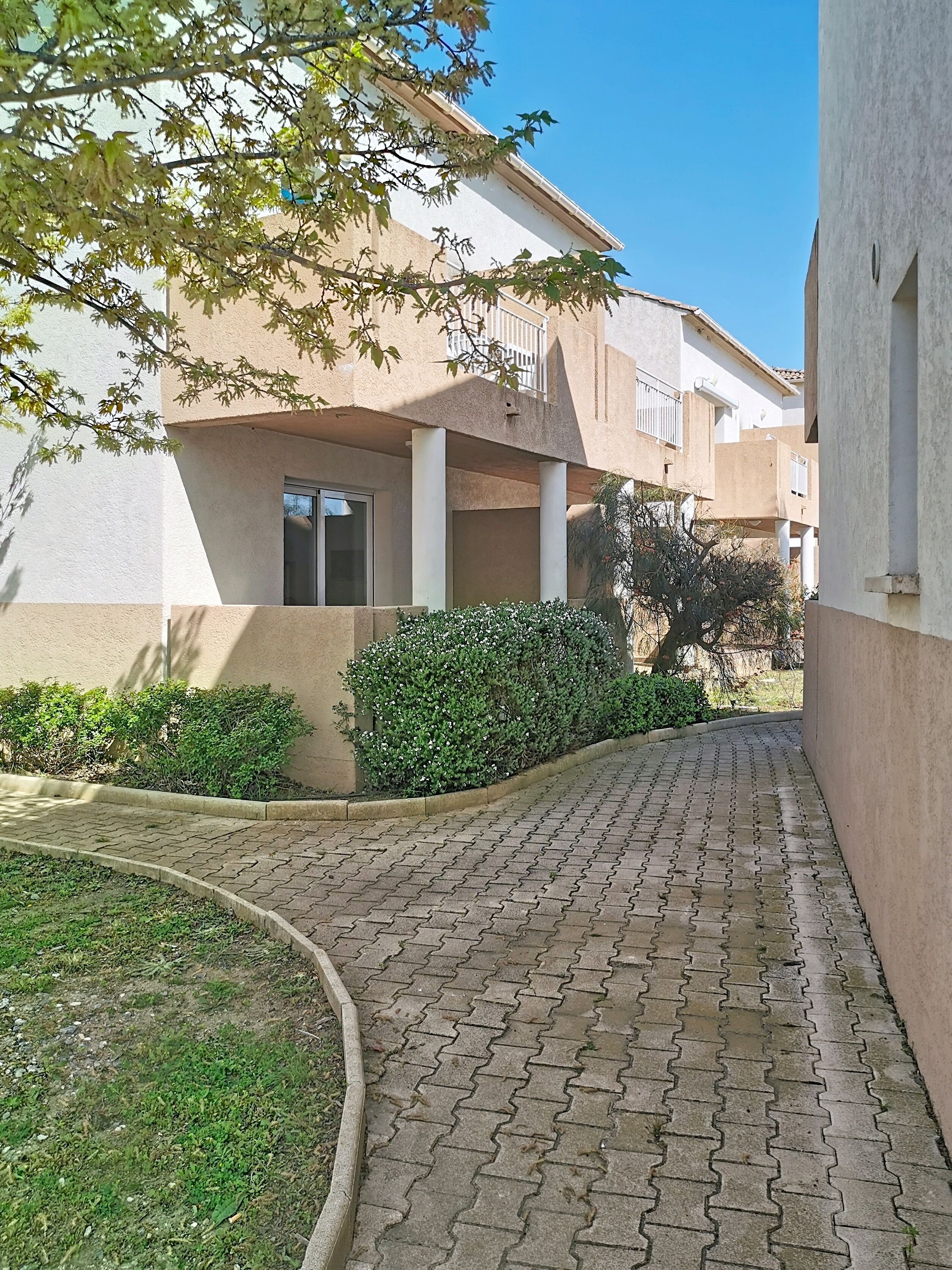 A paved pathway leading to a building with a balcony, surrounded by greenery and a clear sky.