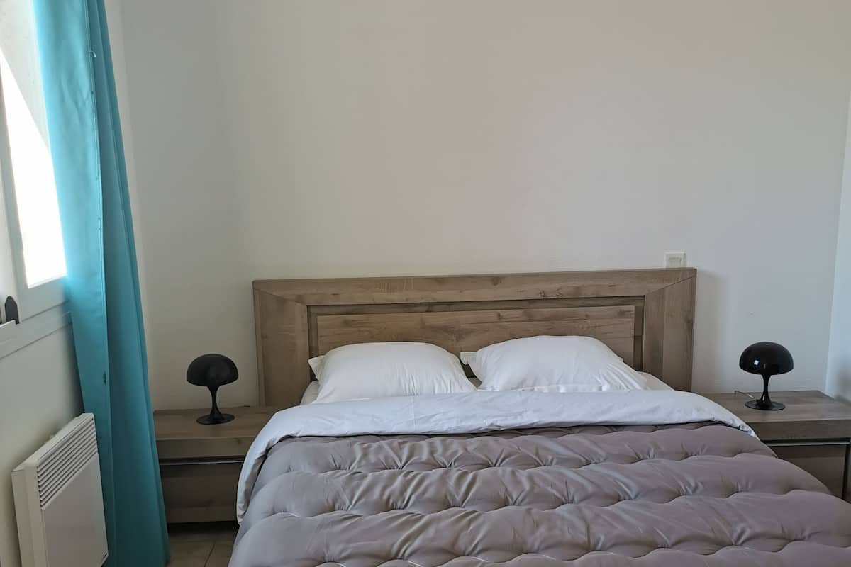 Une chambre à coucher avec un lit, une table de chevet, une lampe et une fenêtre avec des rideaux.