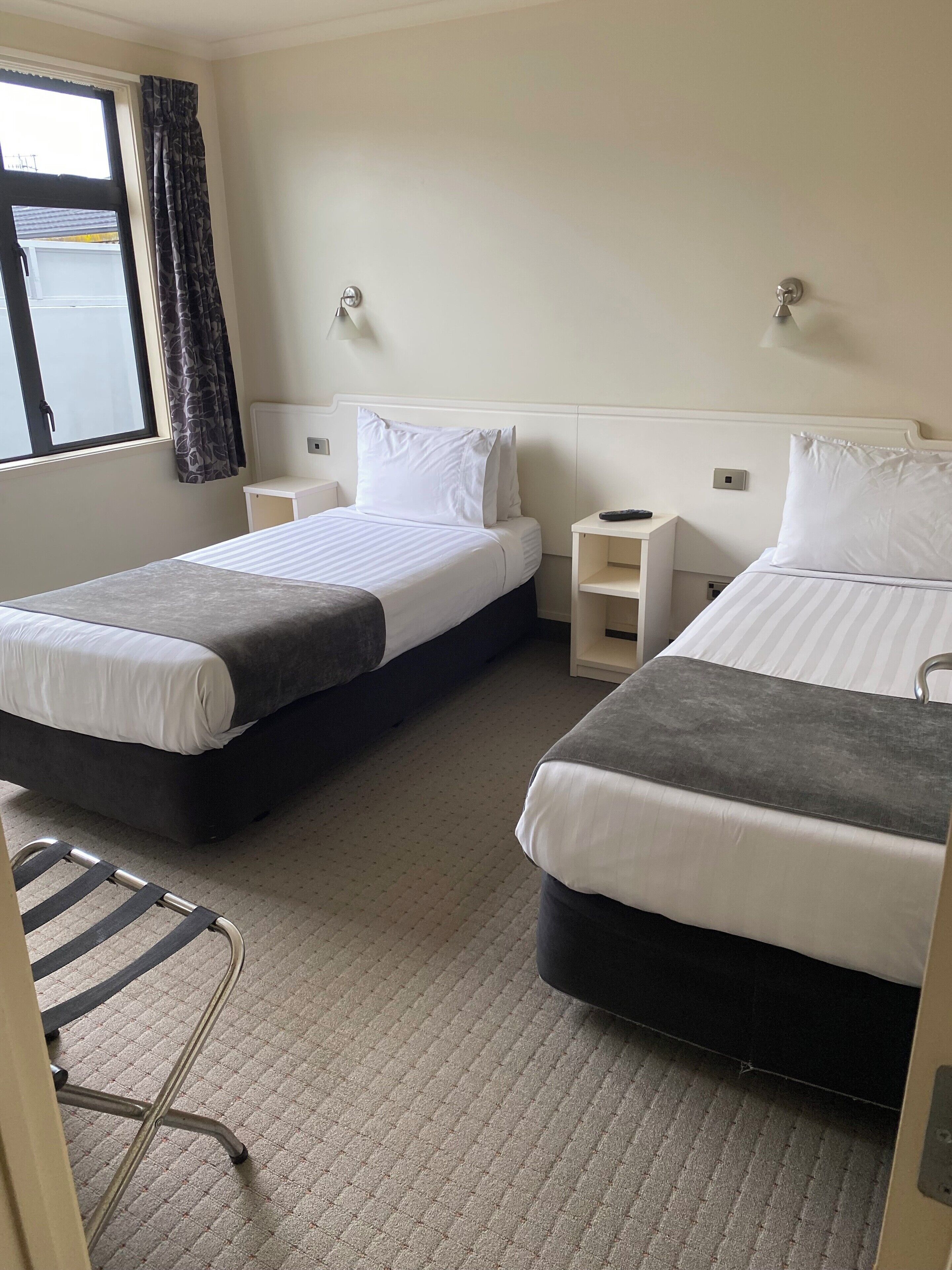 A hotel room with two single beds, a small table, a chair, and a window with curtains.