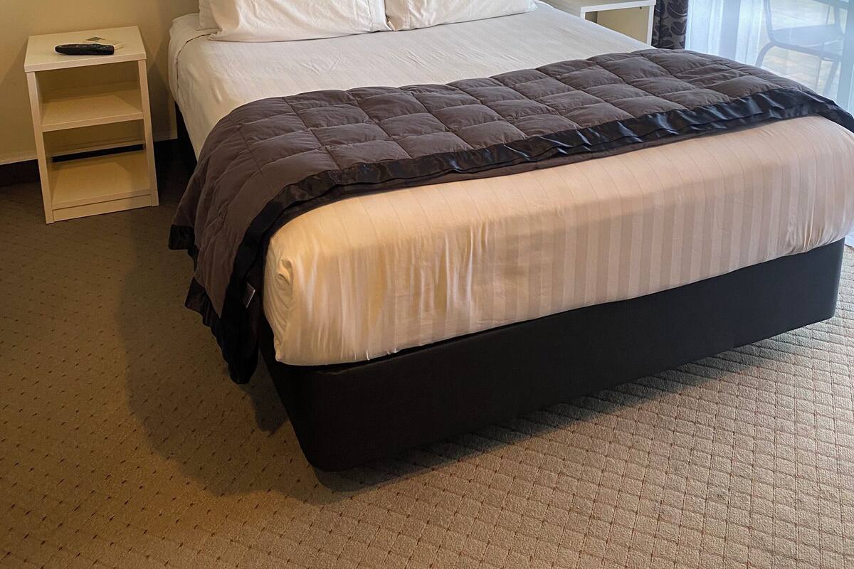 A hotel room with a large bed, a bedside table, a nightstand, a window with curtains, and a wall-mounted light fixture.