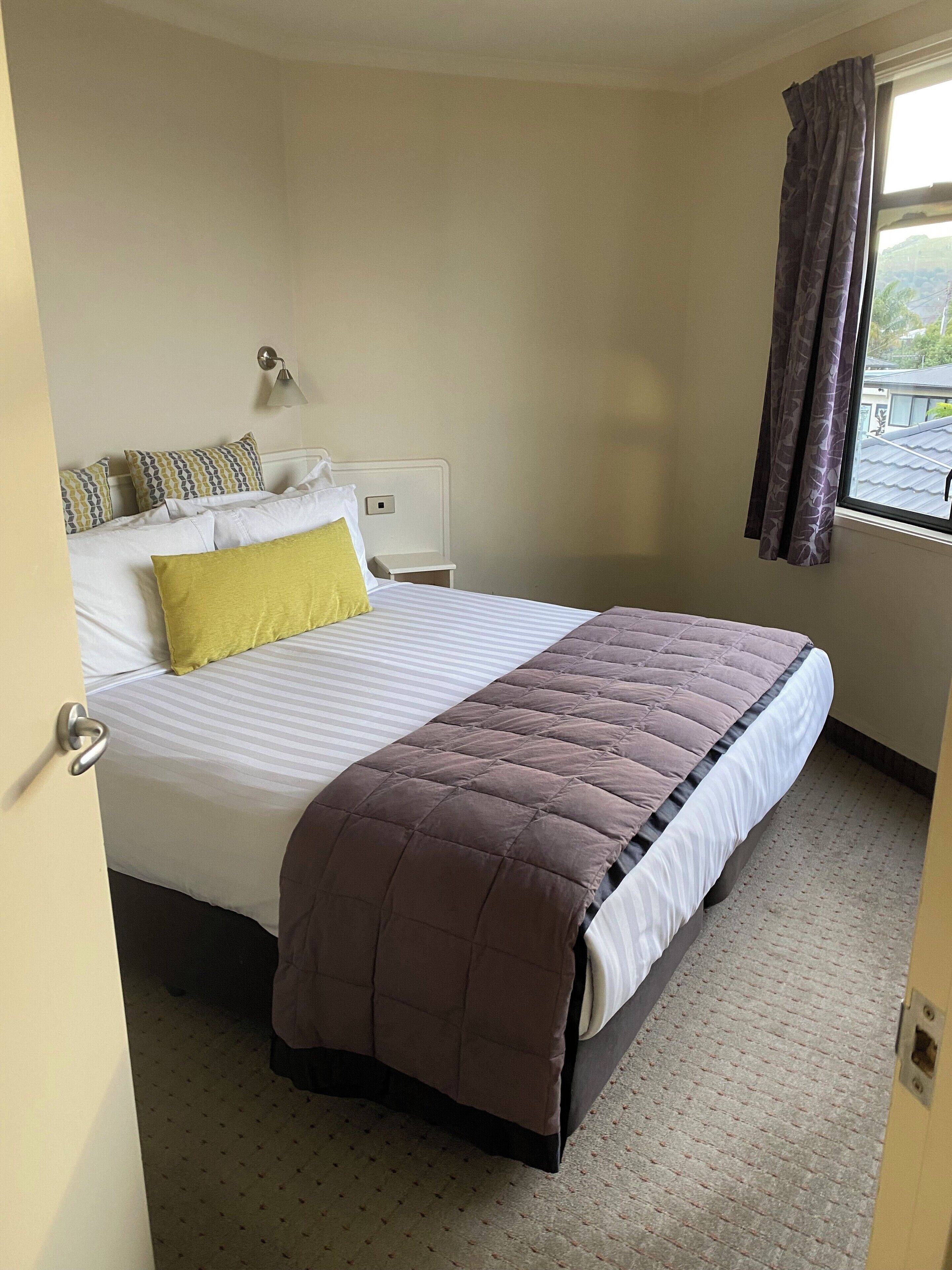 A neatly made bed with a brown duvet, white pillows, and a yellow accent pillow. There is a bedside table with a lamp, a window with curtains, and a view of the outside.