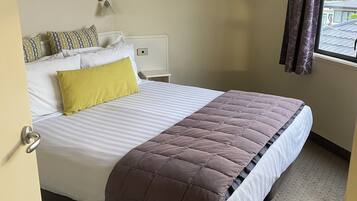 A neatly made bed with a brown duvet, white pillows, and a yellow accent pillow. There is a bedside table with a lamp, a window with curtains, and a view of the outside.