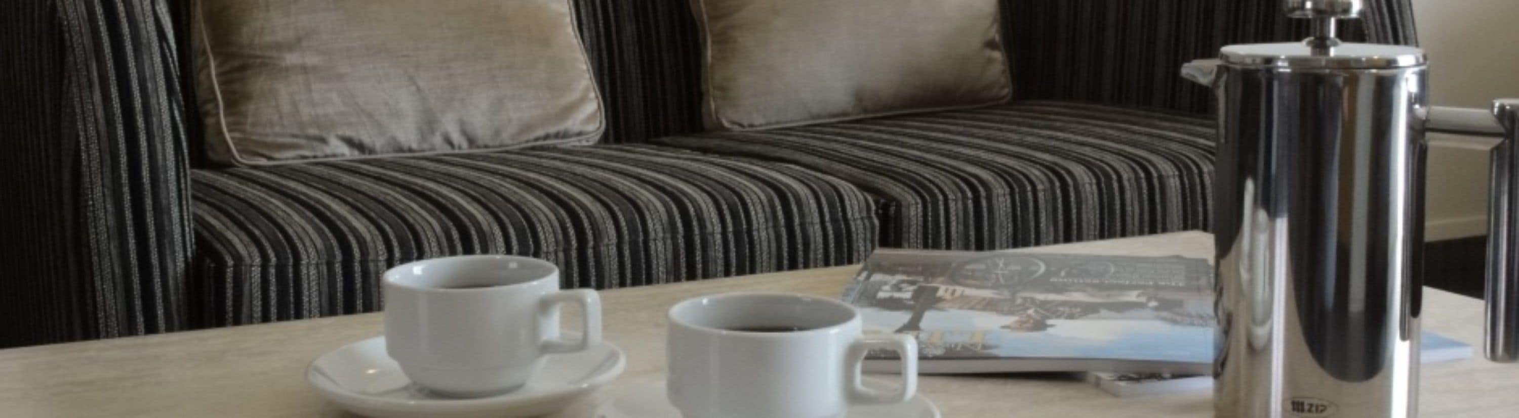 A modern room with a striped sofa, two white cups on a saucer, and a silver coffee pot.