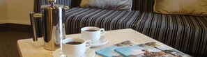 A hotel room with a bed, a coffee maker, two cups of coffee, and a magazine on a table.