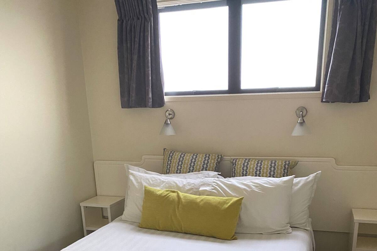 A neatly made bed with a grey duvet and white pillows, a bedside table, a lamp, and a window with curtains.