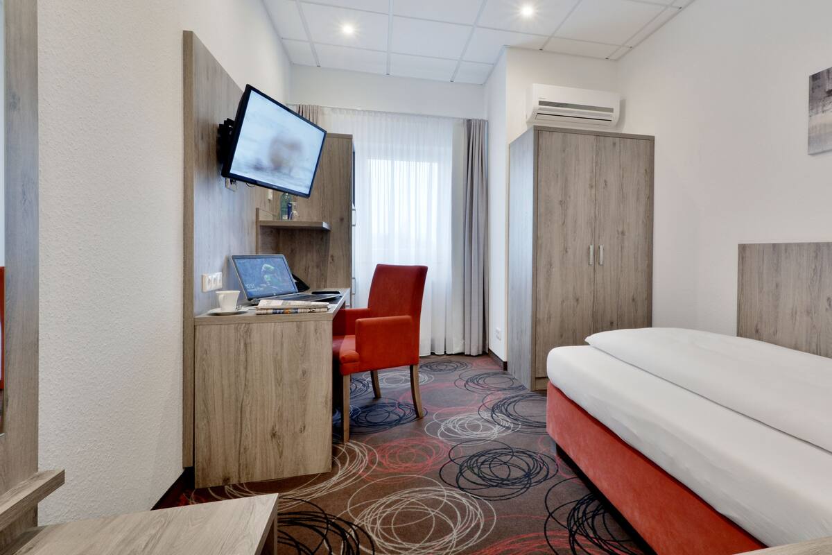 A hotel room with a bed, a desk with a laptop, a chair, a TV mounted on the wall, and an air conditioning unit.