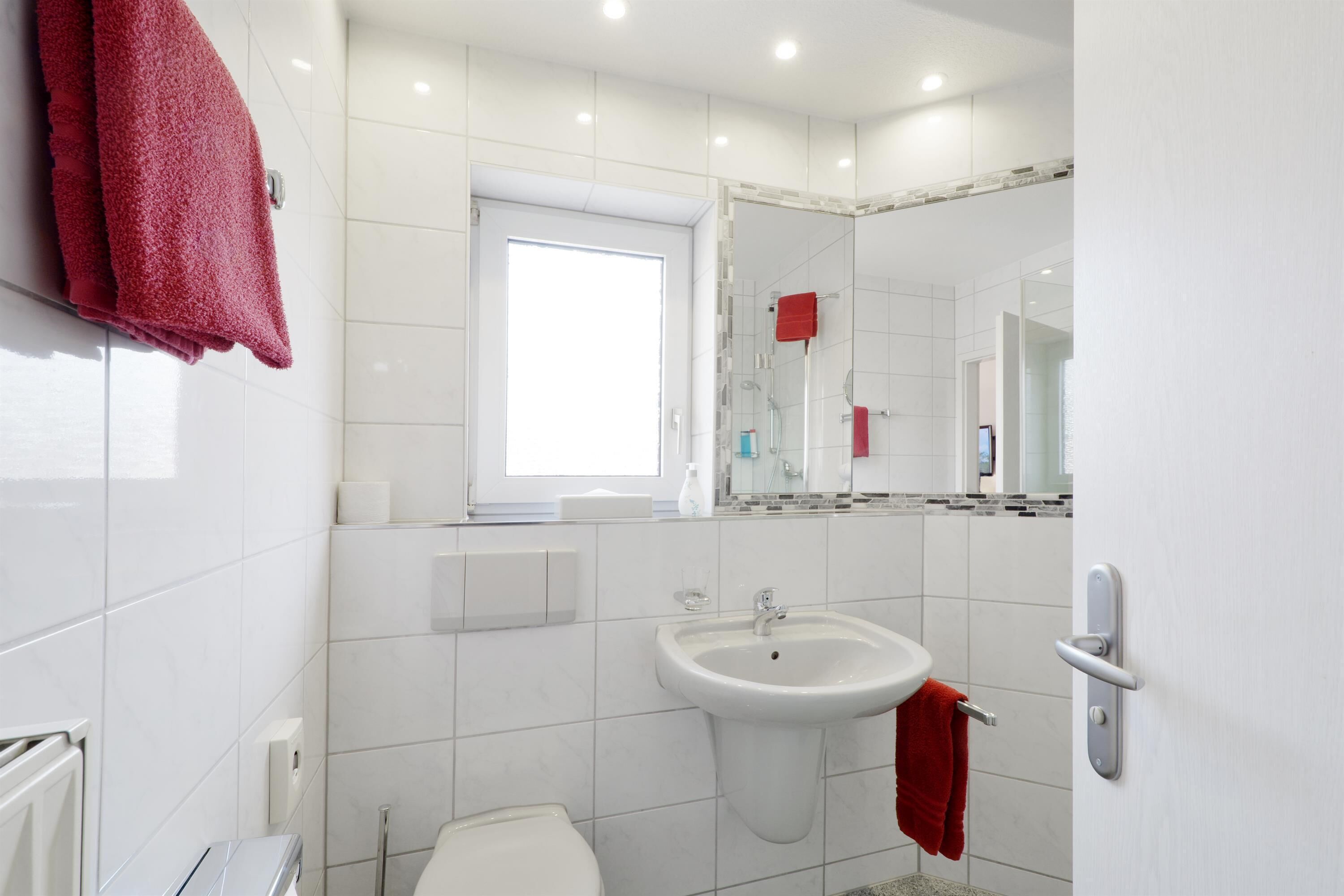 Ein modernes Badezimmer mit weißen Fliesen, einem Waschbecken, einer Toilette und einem Spiegel.