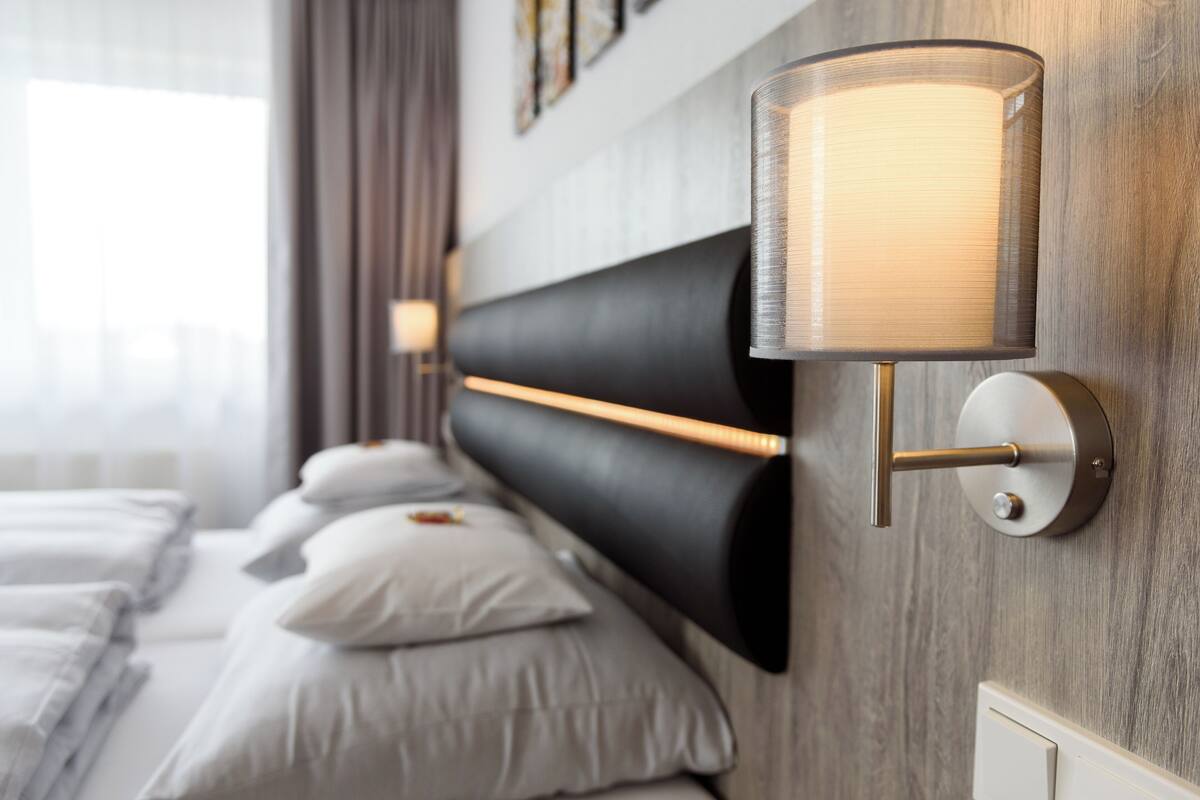 A modern hotel room with a large bed, a headboard with integrated lighting, and a wall-mounted lamp.
