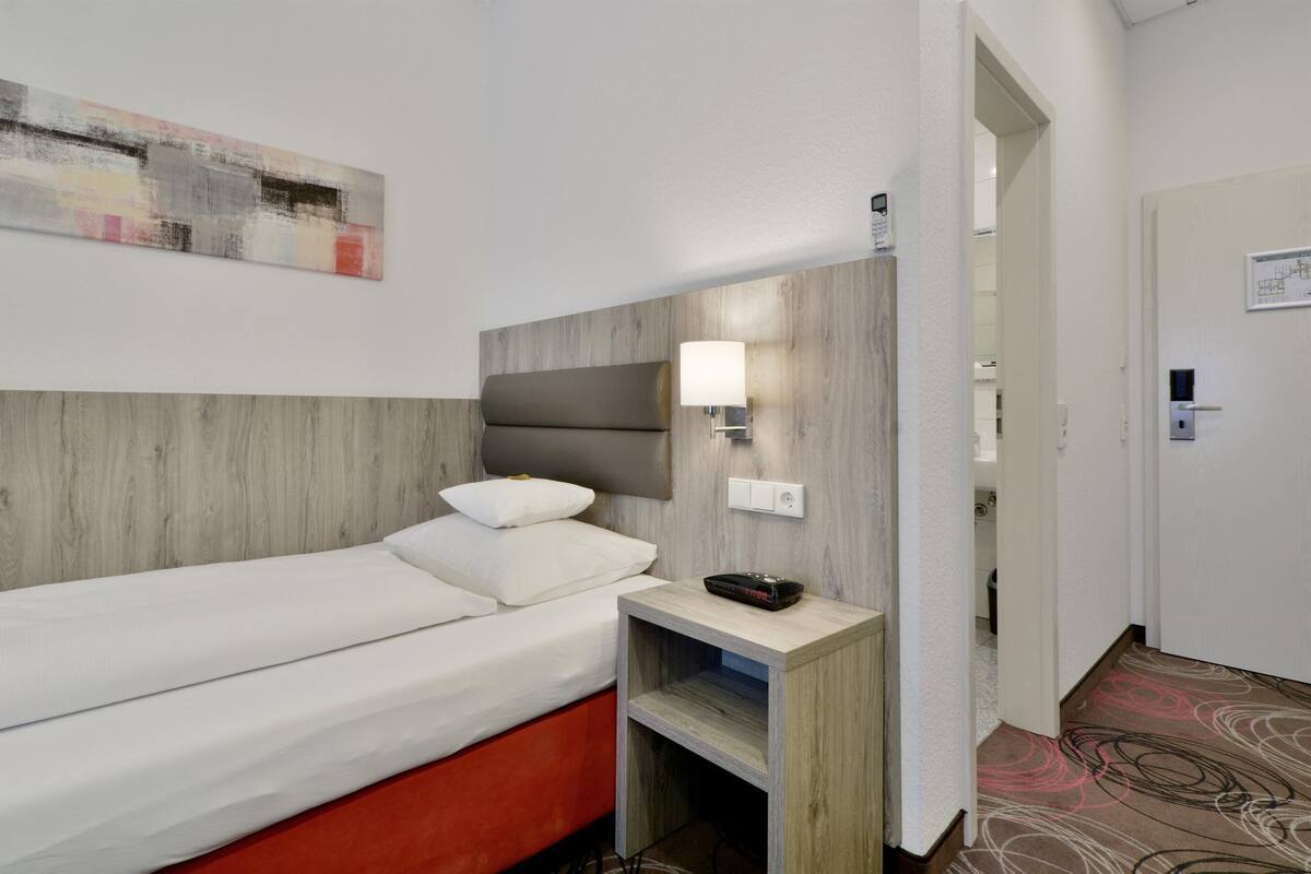A hotel room with a bed, a nightstand, a wall-mounted lamp, and a painting.