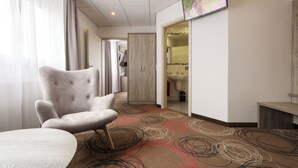 A hotel room with a large armchair, a round table, a TV mounted on the wall, and a carpet with circular patterns. - Hotel Engelhorn (Leimen)