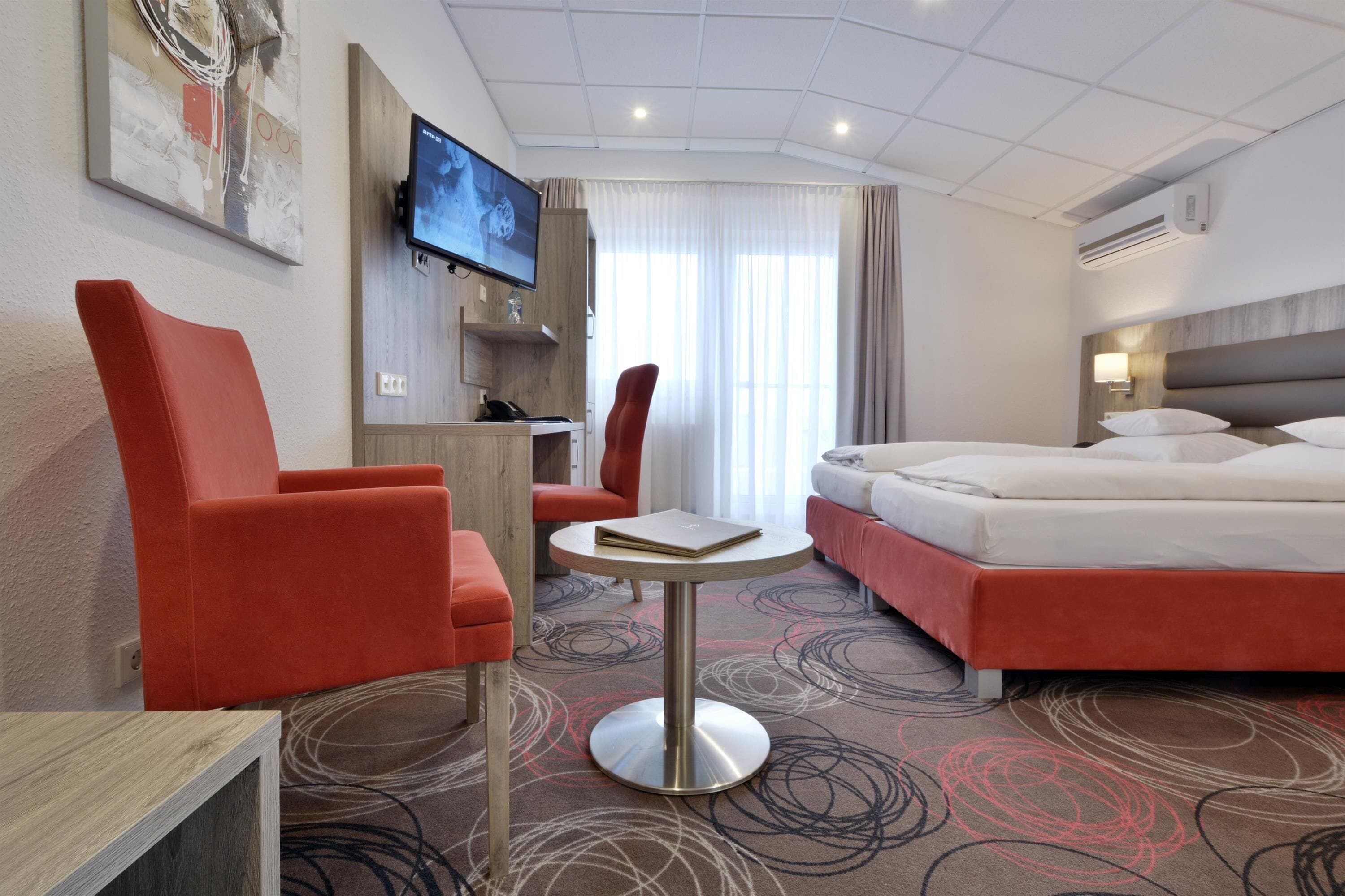 A hotel room with a bed, two red armchairs, a small round table, a TV mounted on the wall, and a desk with a chair.
