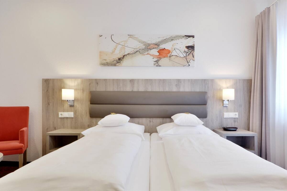 A hotel room with two beds, a red armchair, a bedside table, and a wall-mounted abstract painting.