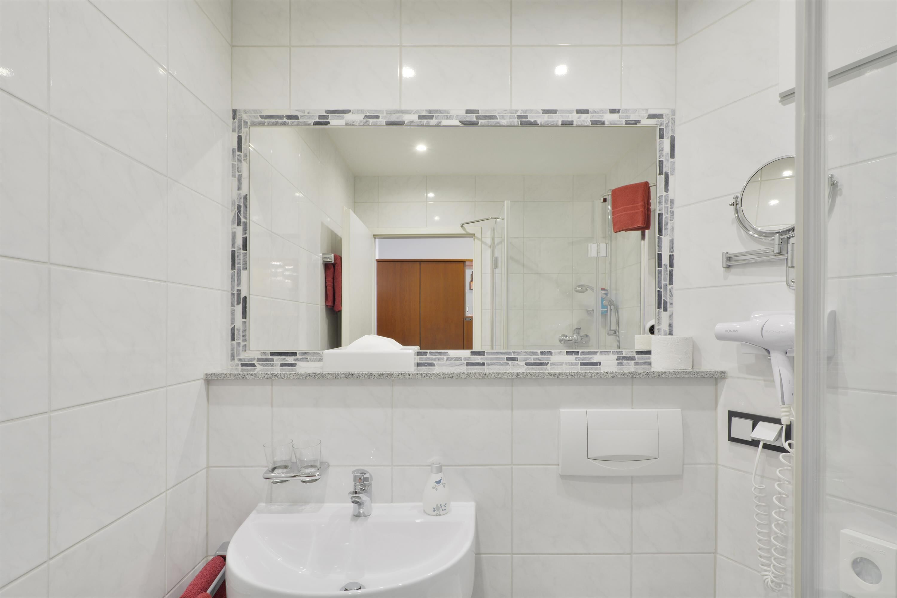 A modern bathroom with a white sink, a mirror with a mosaic border, and a hairdryer on the wall.