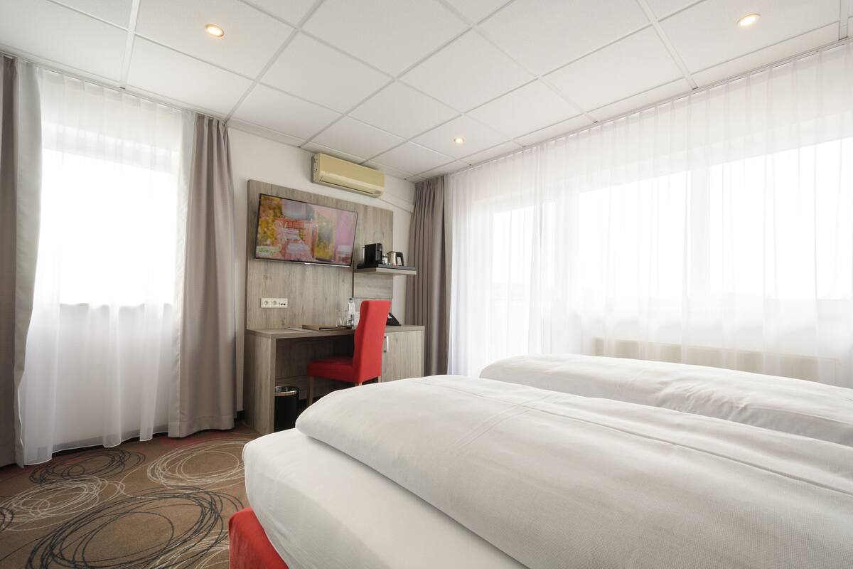 A hotel room with a large bed, a desk with a chair, a painting on the wall, and a window with curtains.