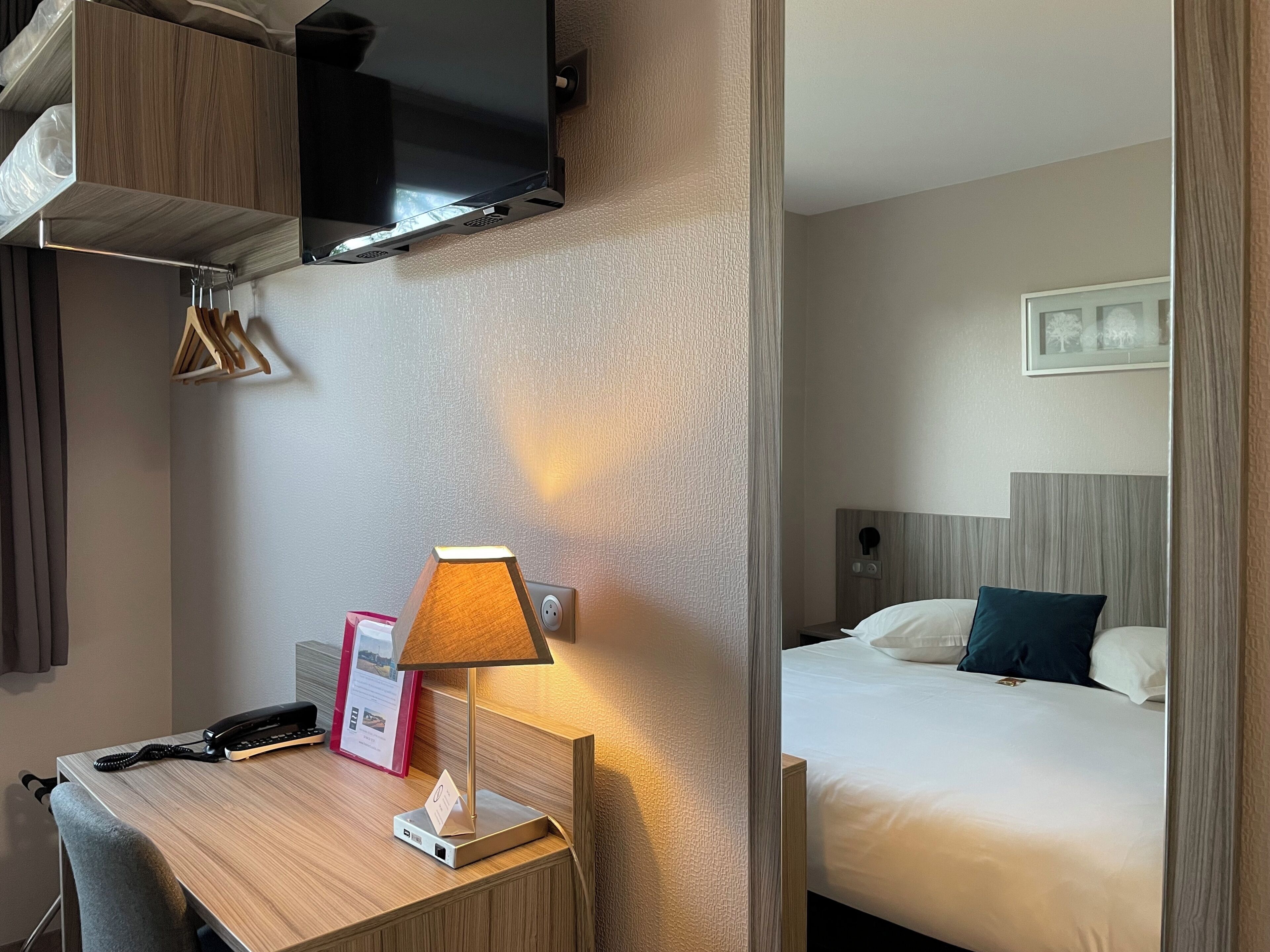 A hotel room with a bed, a desk with a lamp, a TV mounted on the wall, and a coat rack.