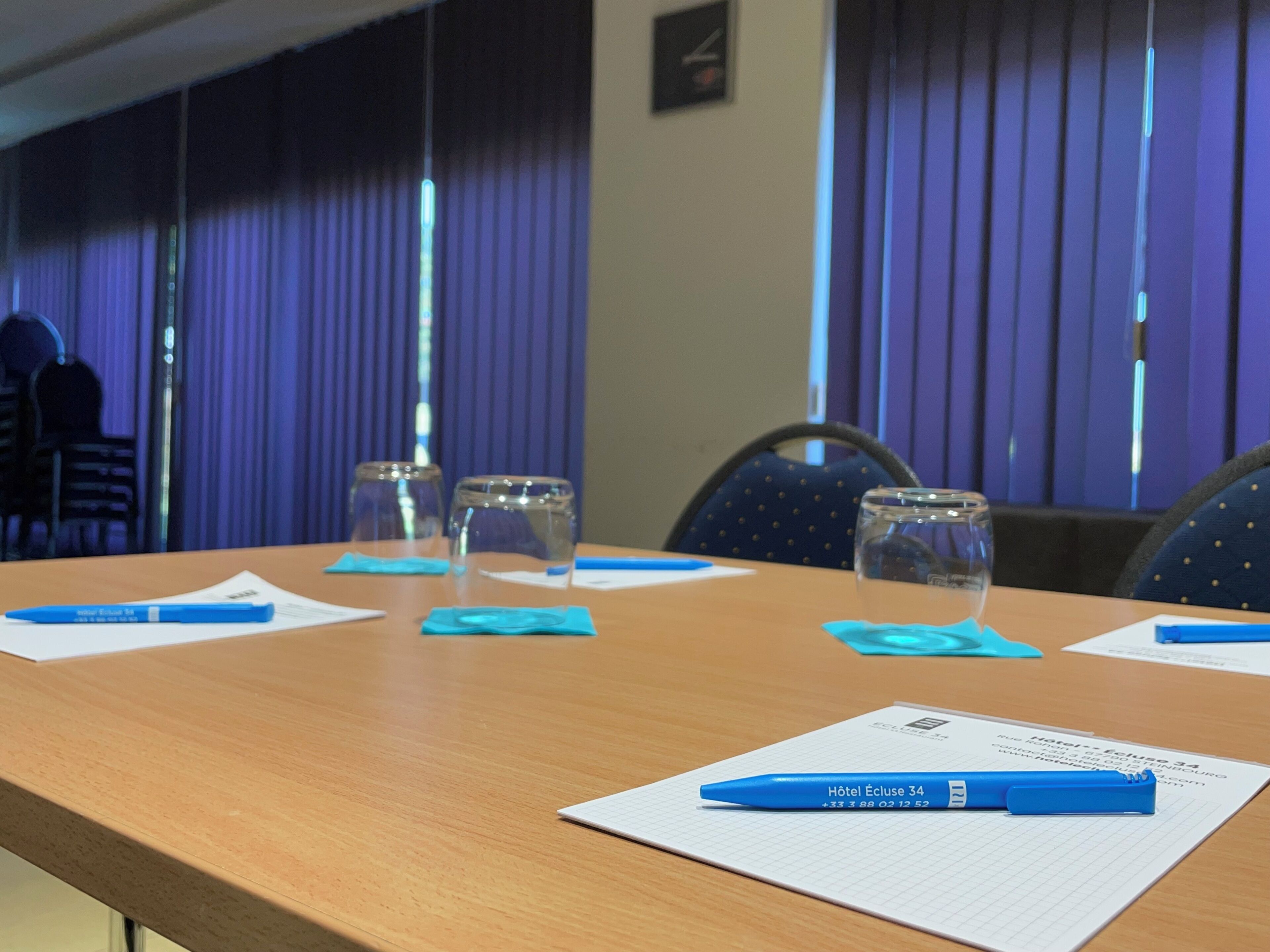 Una sala de conferencias con una mesa de madera, vasos de vidrio con agua y bolígrafos azules sobre blocs de notas.