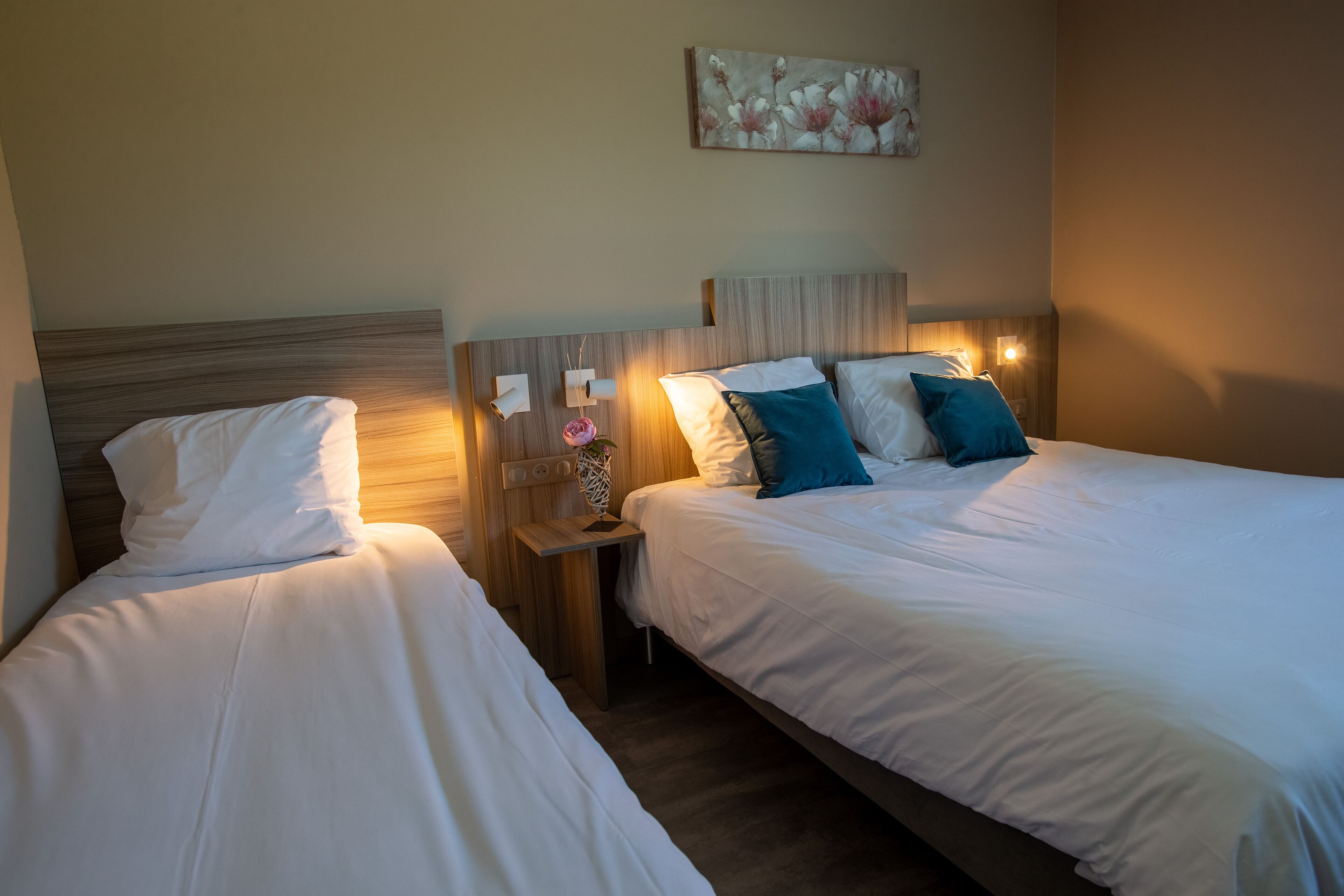 A hotel room with two beds, each with white bedding and decorative pillows. There is a nightstand with a lamp and a vase with flowers. A painting is visible on the wall above the beds.