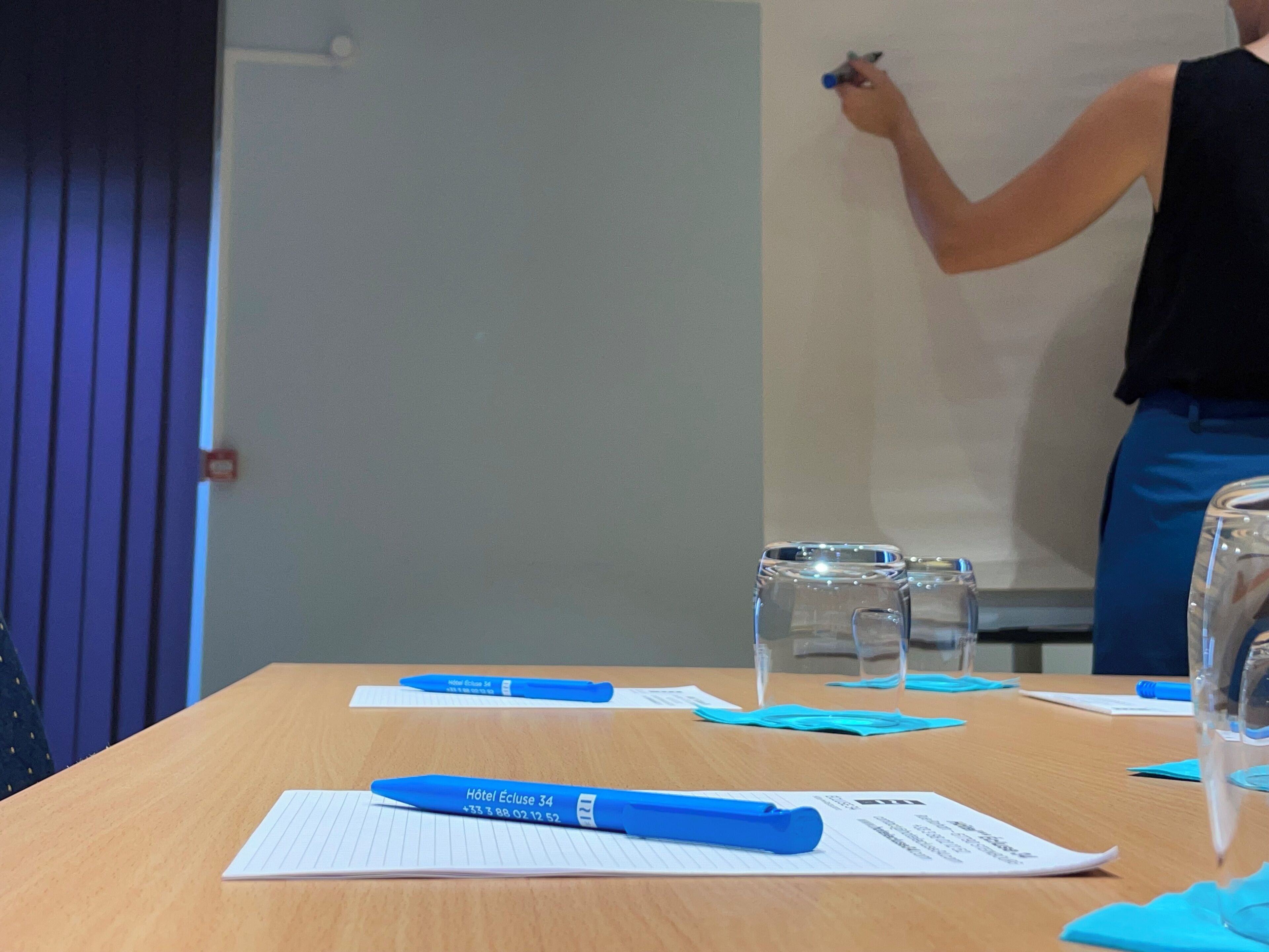 A meeting room with a whiteboard, blue pens, papers, and a glass of water on the table.