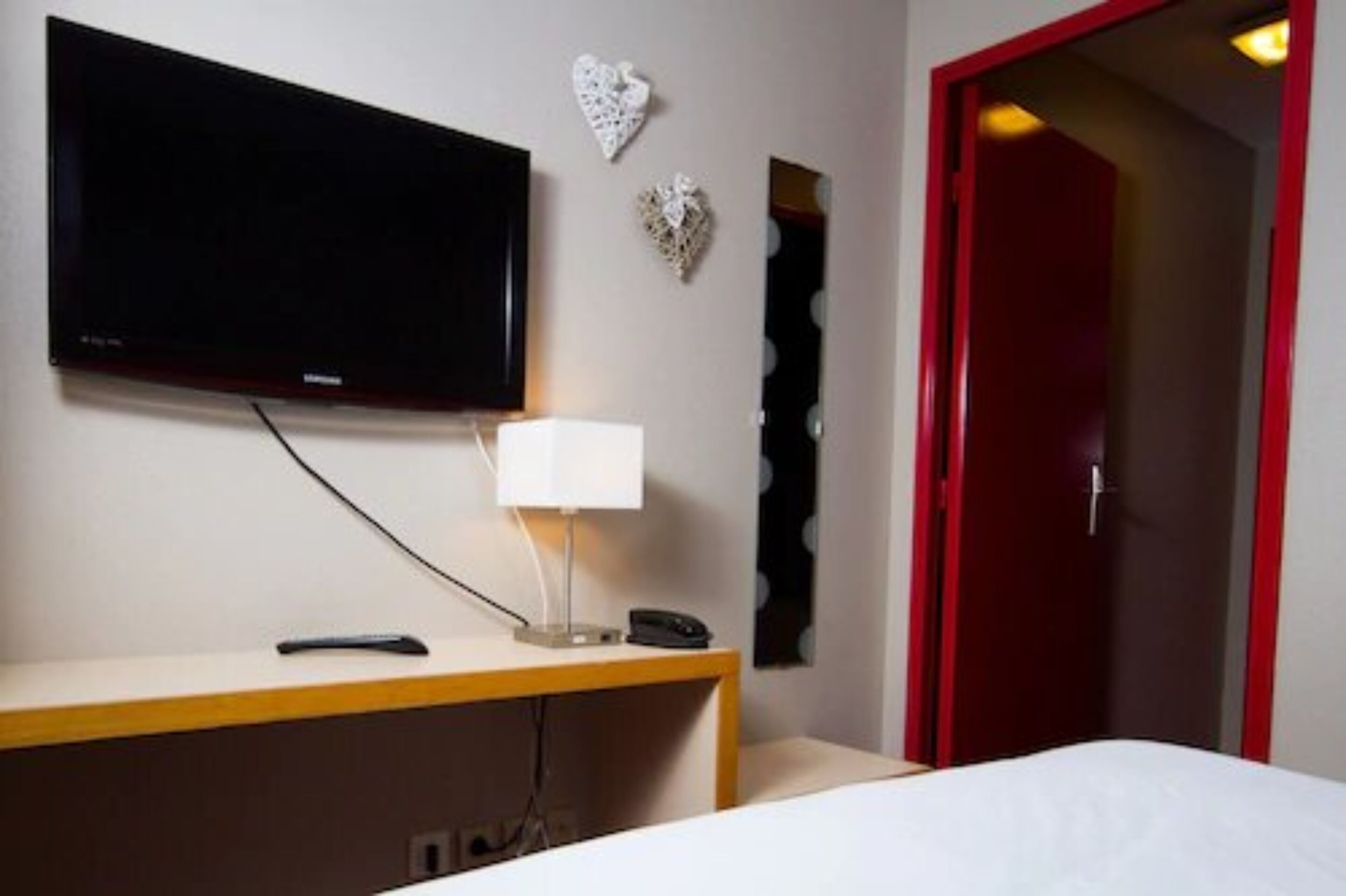 A hotel room with a flat-screen TV mounted on the wall, a bedside table with a lamp, a phone, and a bed.