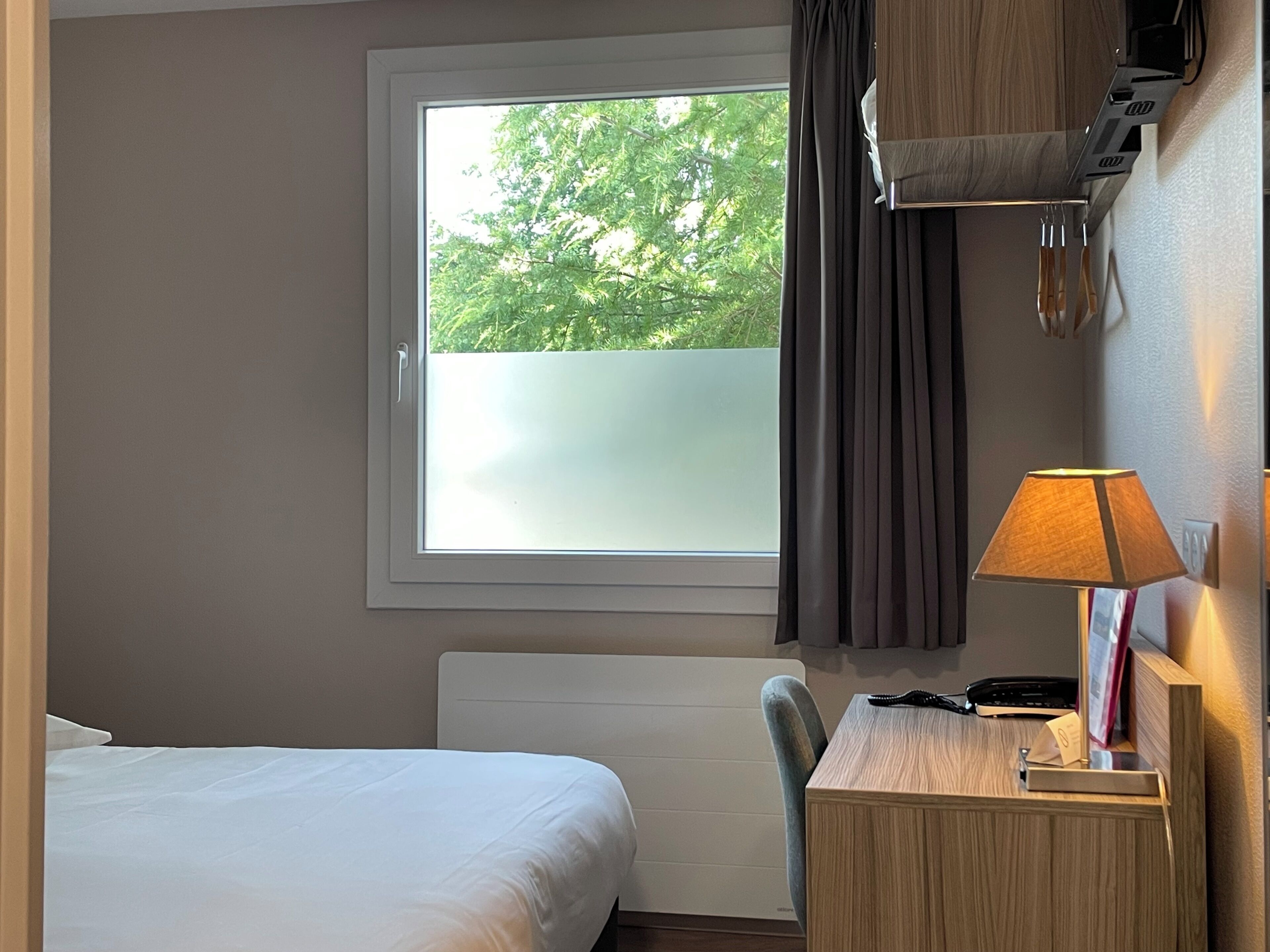 Habitación de hotel con cama, escritorio con lámpara, silla, ventana con cortinas y vista a árboles.