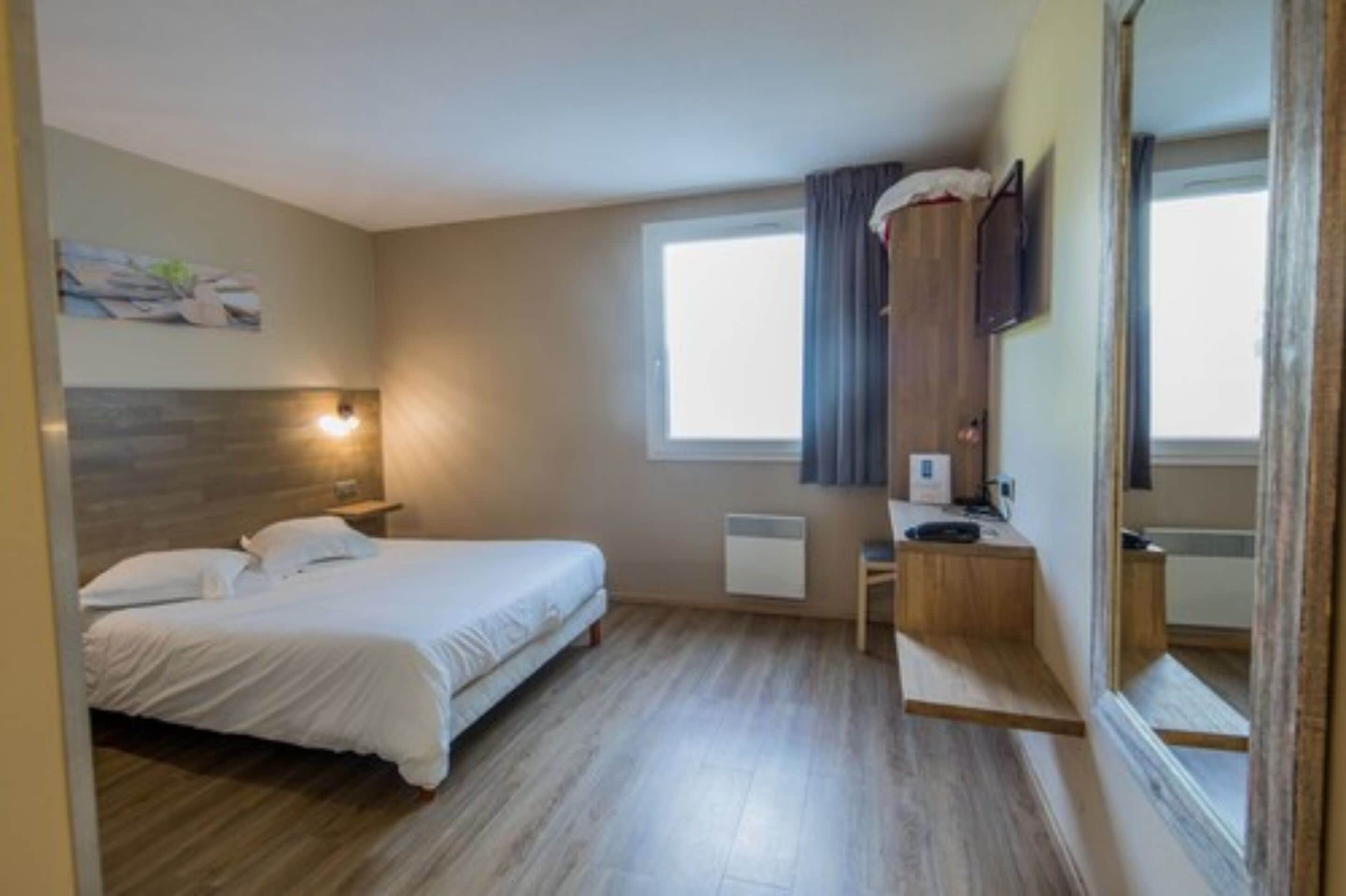 A bedroom with a bed, bedside table, lamp, window, and a mirror.