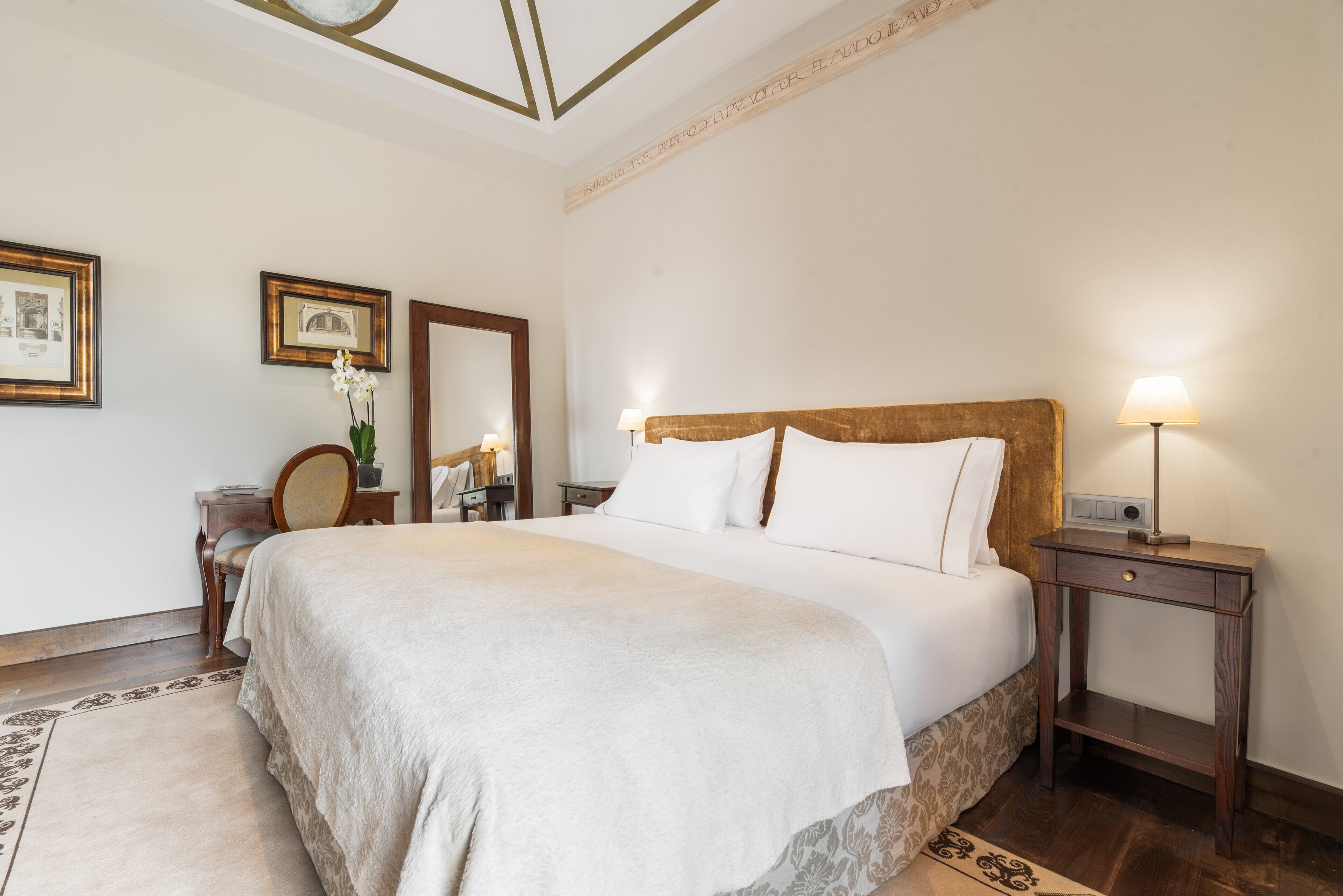 A neatly arranged bedroom with a large bed, bedside tables, a mirror, and framed pictures on the wall.