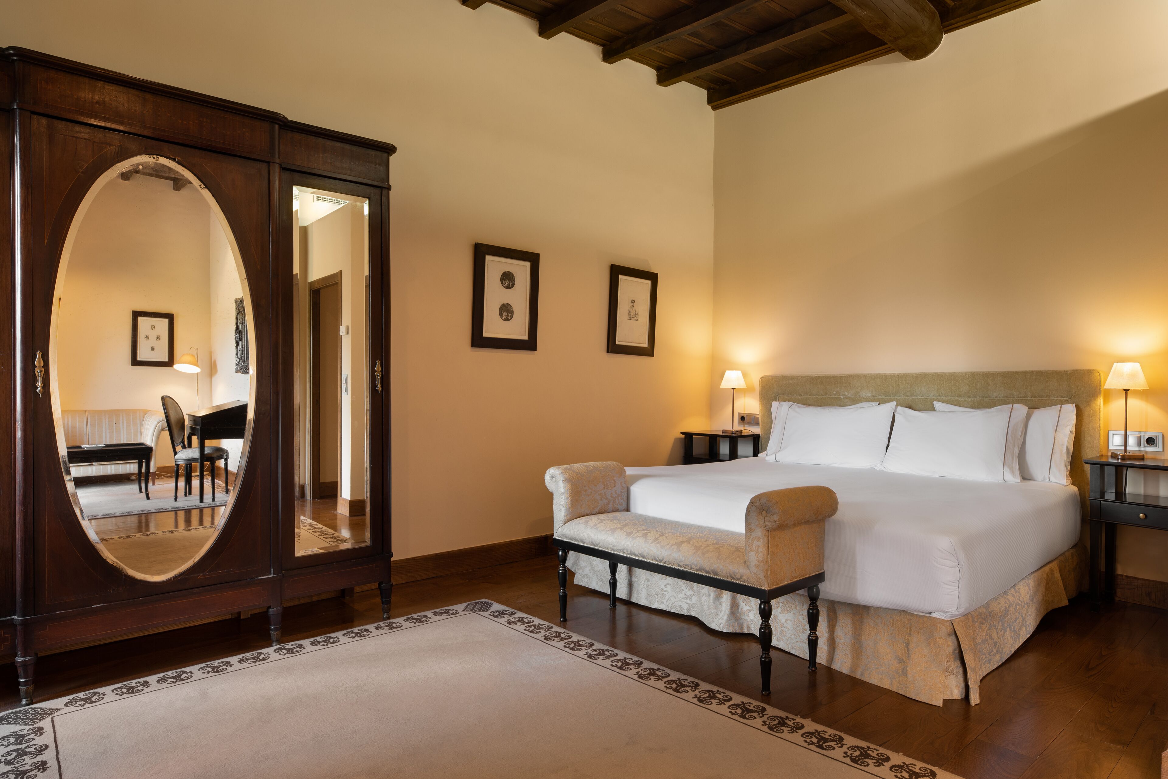 A bedroom with a large mirror, a bed with white linens, a bench, and a wooden wardrobe.