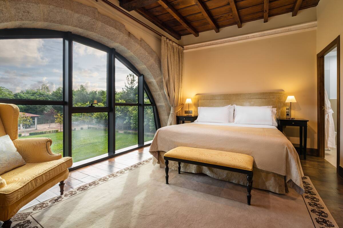Une chambre avec une grande fenêtre, un lit, un banc et une chaise.