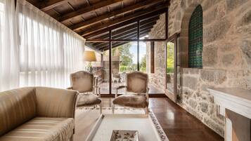 Un cuarto acogedor con pared de piedra, techo de madera y un gran ventanal. Hay un sofá, dos sillones y una mesita con una lámpara.