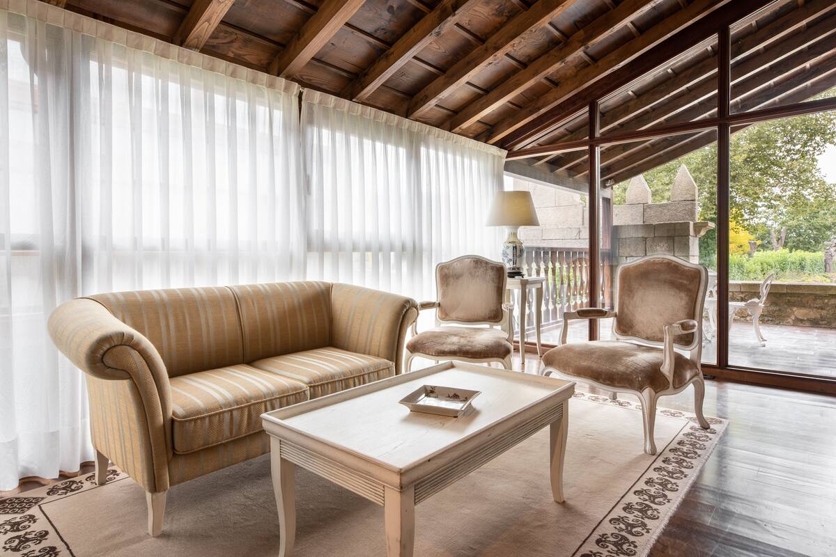 Une pièce avec un plafond en bois, un canapé beige, deux fauteuils, une petite table et une vue sur l’extérieur grâce à de grandes fenêtres.