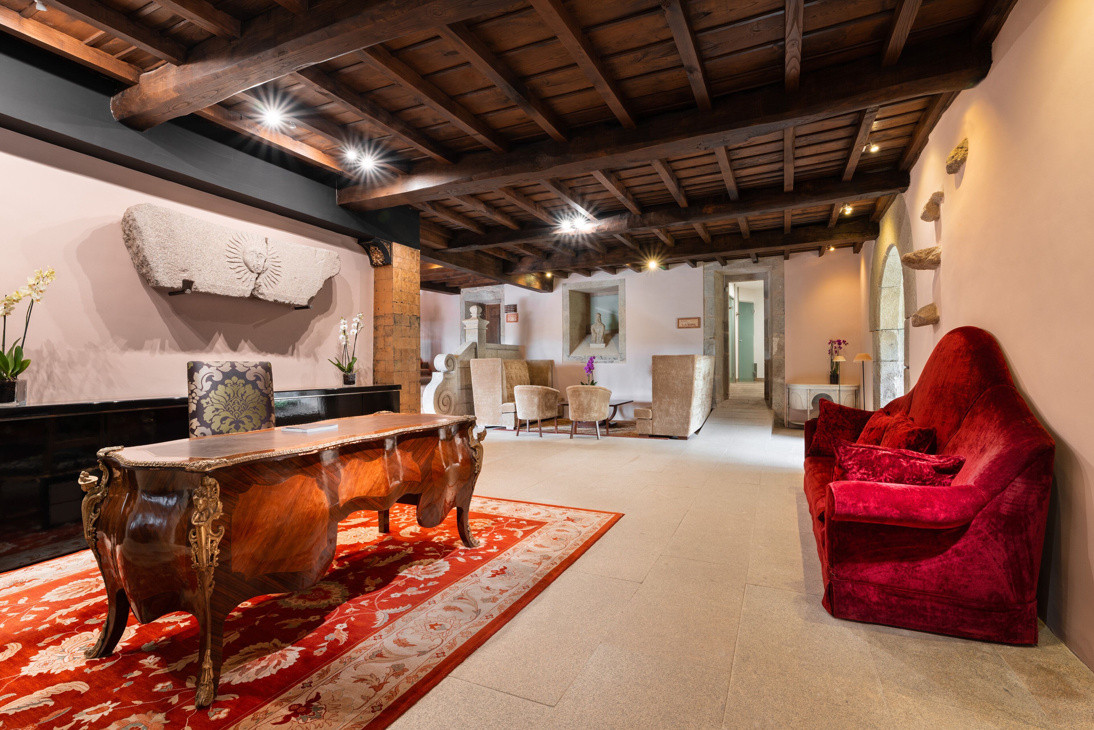 A spacious room with a wooden ceiling, a large wooden table, a red velvet sofa, and a red carpet.