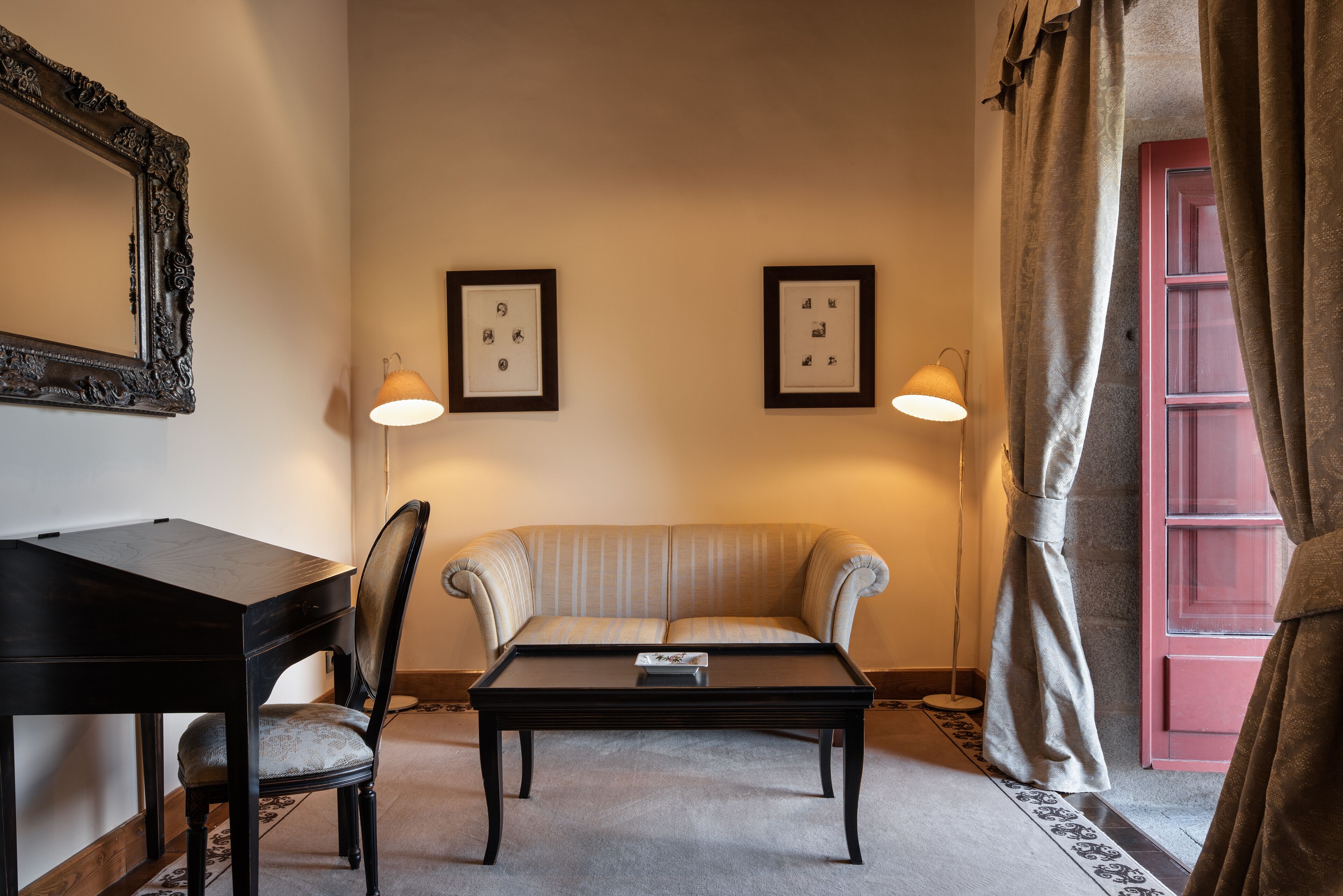 A room with a dark wooden desk, a cushioned sofa, a small table, two framed pictures on the wall, and a lamp.