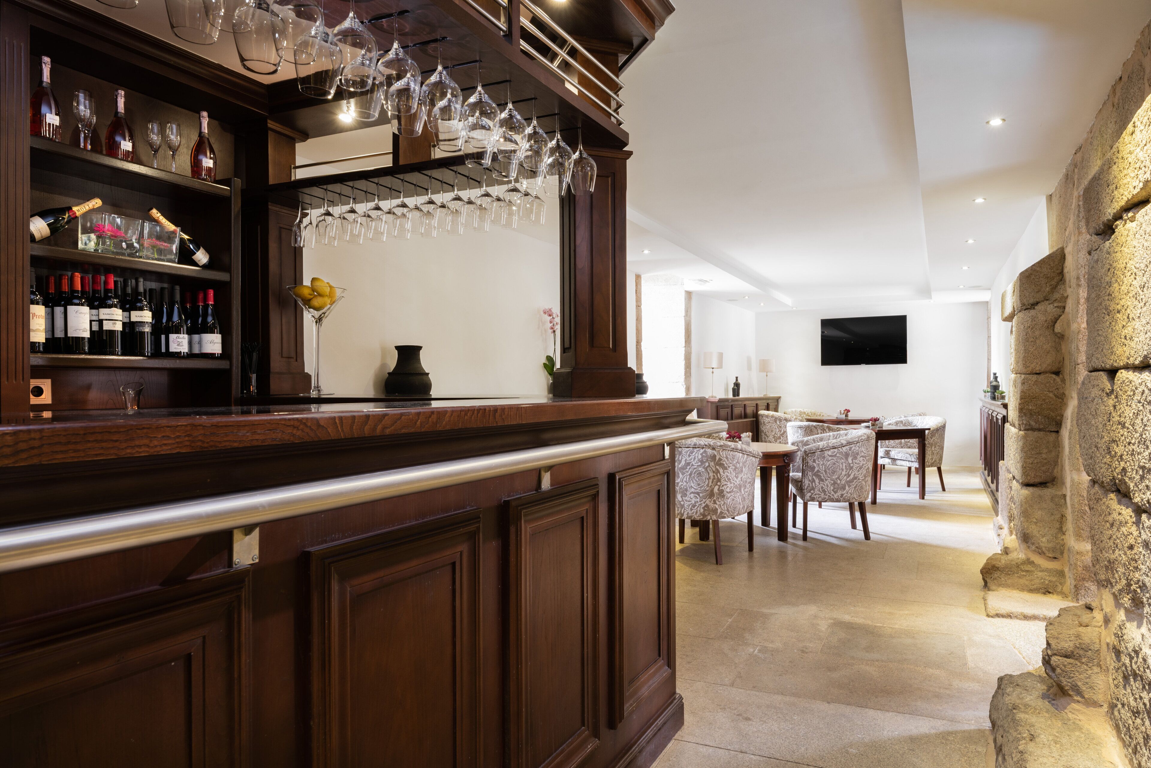 Zona de bar con armarios de madera, botellas de vino y copas. Se observa una pared de piedra y un área de comedor con sillas y una mesa.