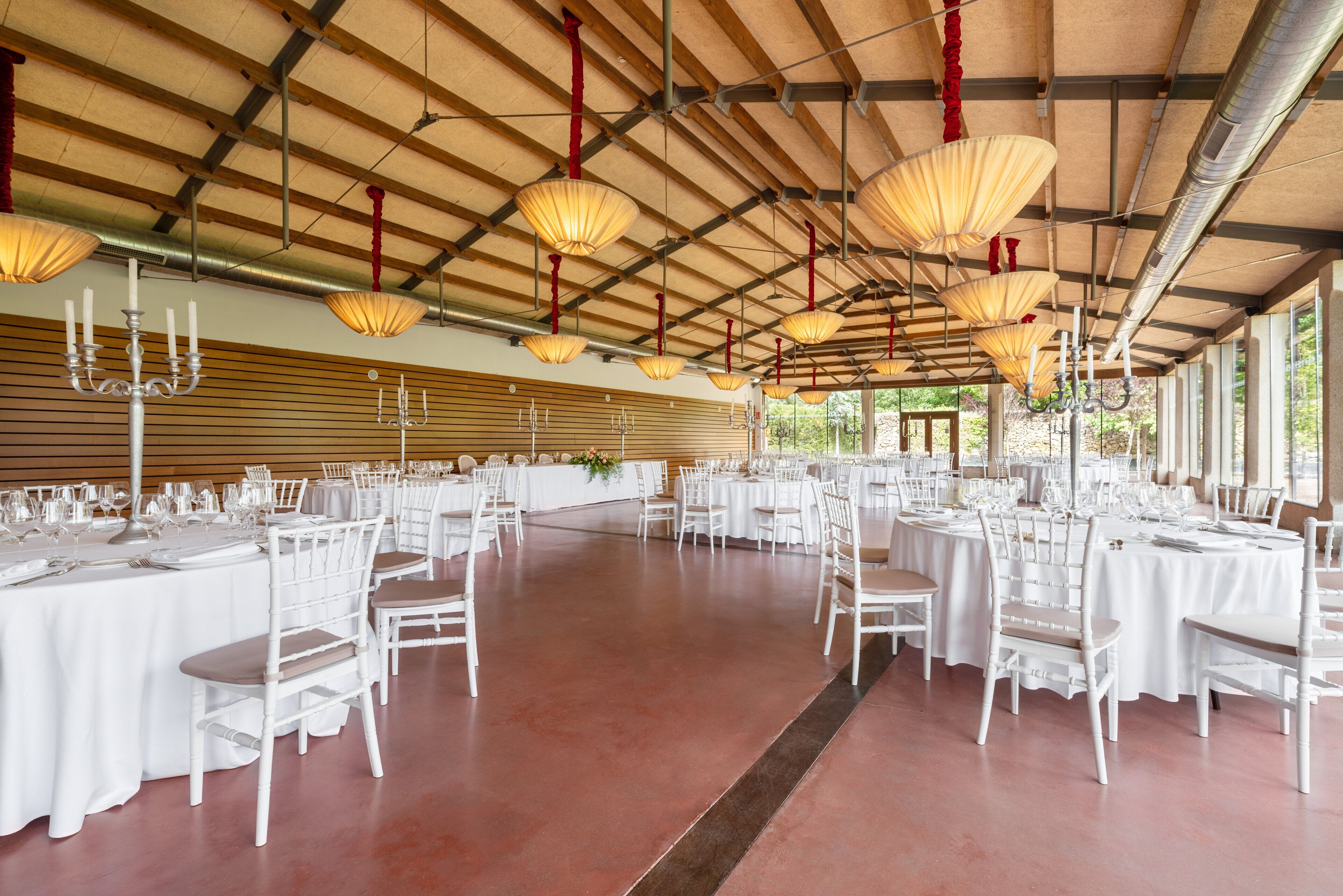 A spacious hall with round tables and white chairs, large hanging lamps, and a high ceiling with visible beams.