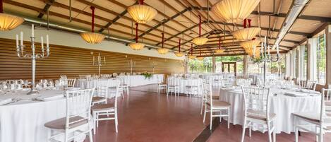 A spacious hall with round tables and white chairs, large hanging lamps, and a high ceiling with visible beams.