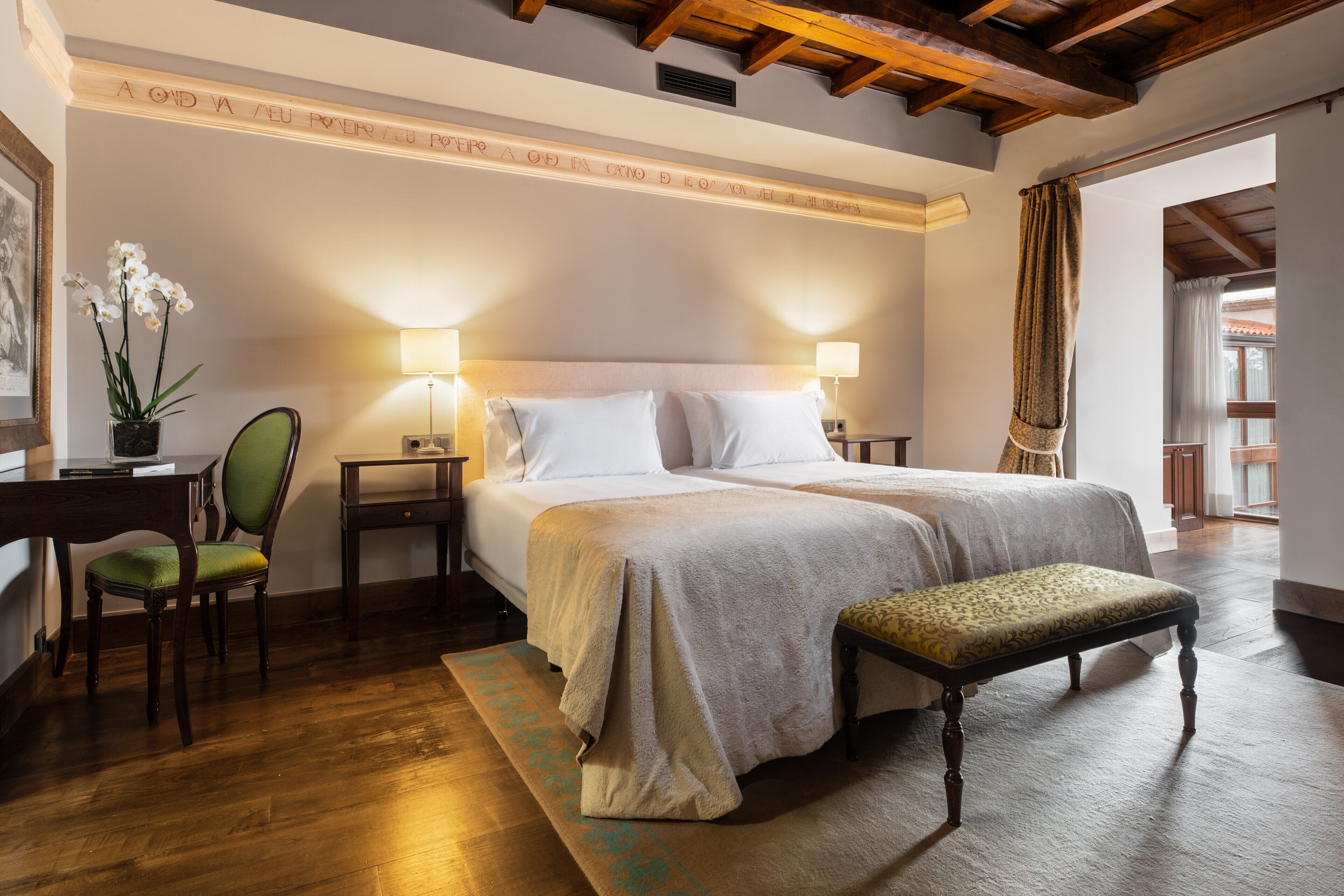 Habitación de hotel con dos camas, un escritorio con silla, un banco y un ventanal grande.