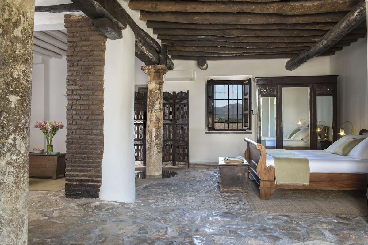 A rustic room with a stone floor, a wooden bed, a large mirror, and a window with bars.