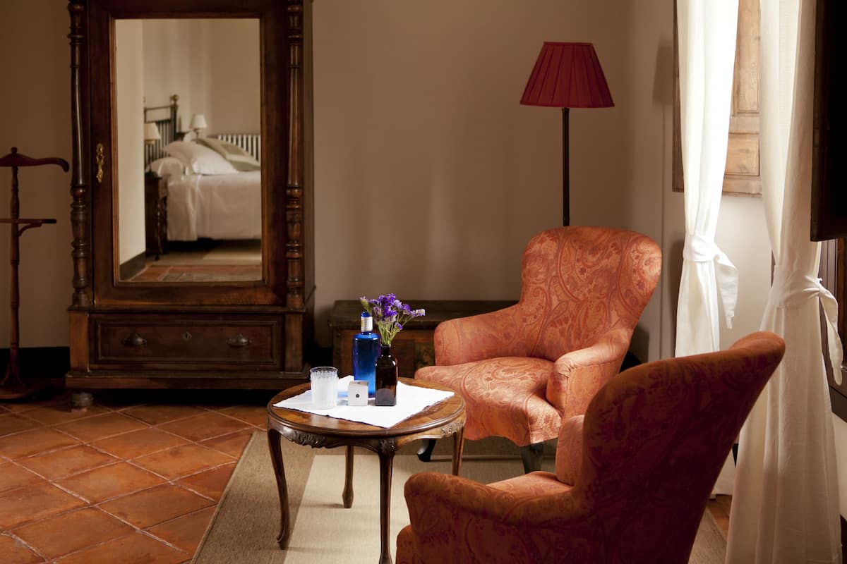 A room with a large mirror, a wooden dresser, an orange patterned armchair, a small round table with a vase and a glass, and a red floor lamp.