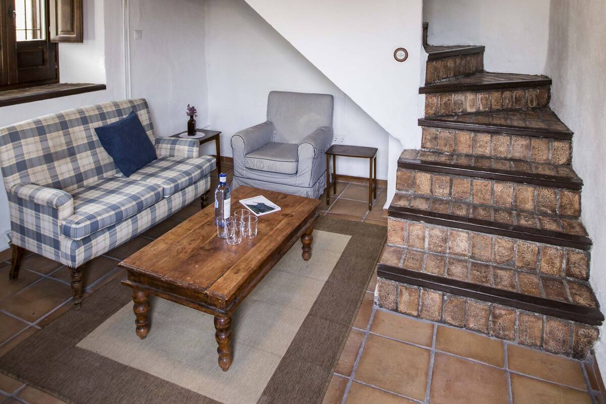A cozy living room with a wooden coffee table, a plaid sofa, a striped armchair, and a brick staircase.