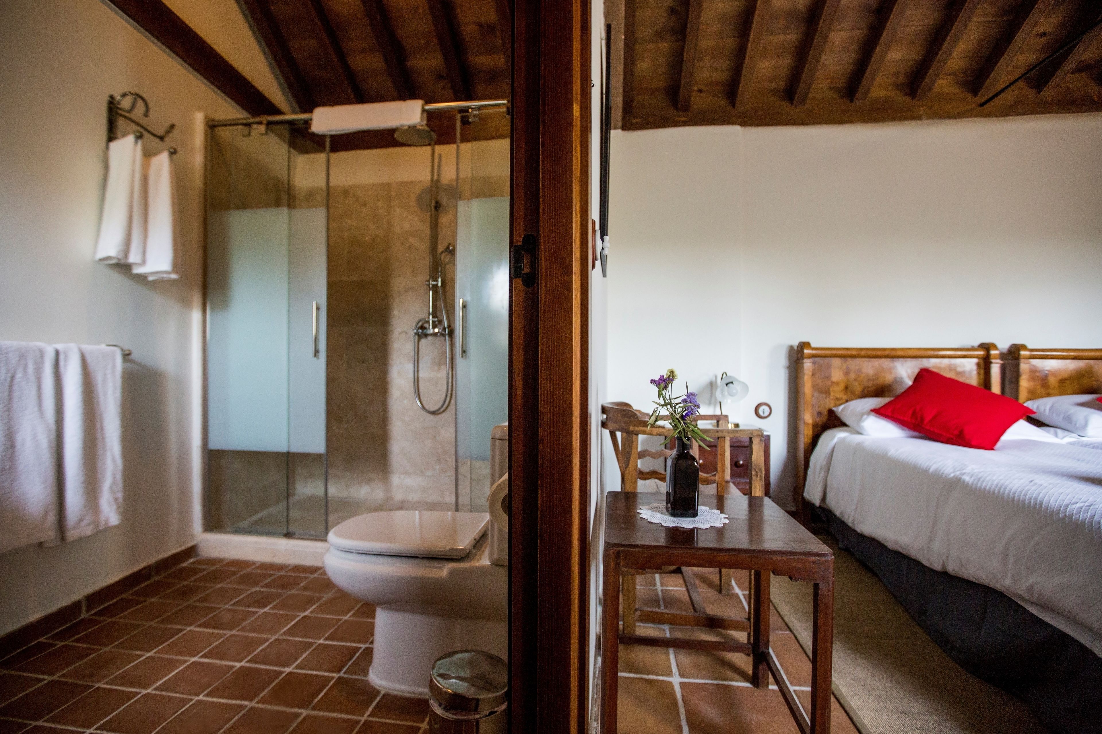 A bathroom with a glass-enclosed shower, a toilet, and a sink. There is a wooden table with a vase of flowers and a chair.