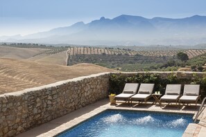 Zona de piscina con muros de piedra y sillones de descanso, con vistas a un viñedo y montañas.