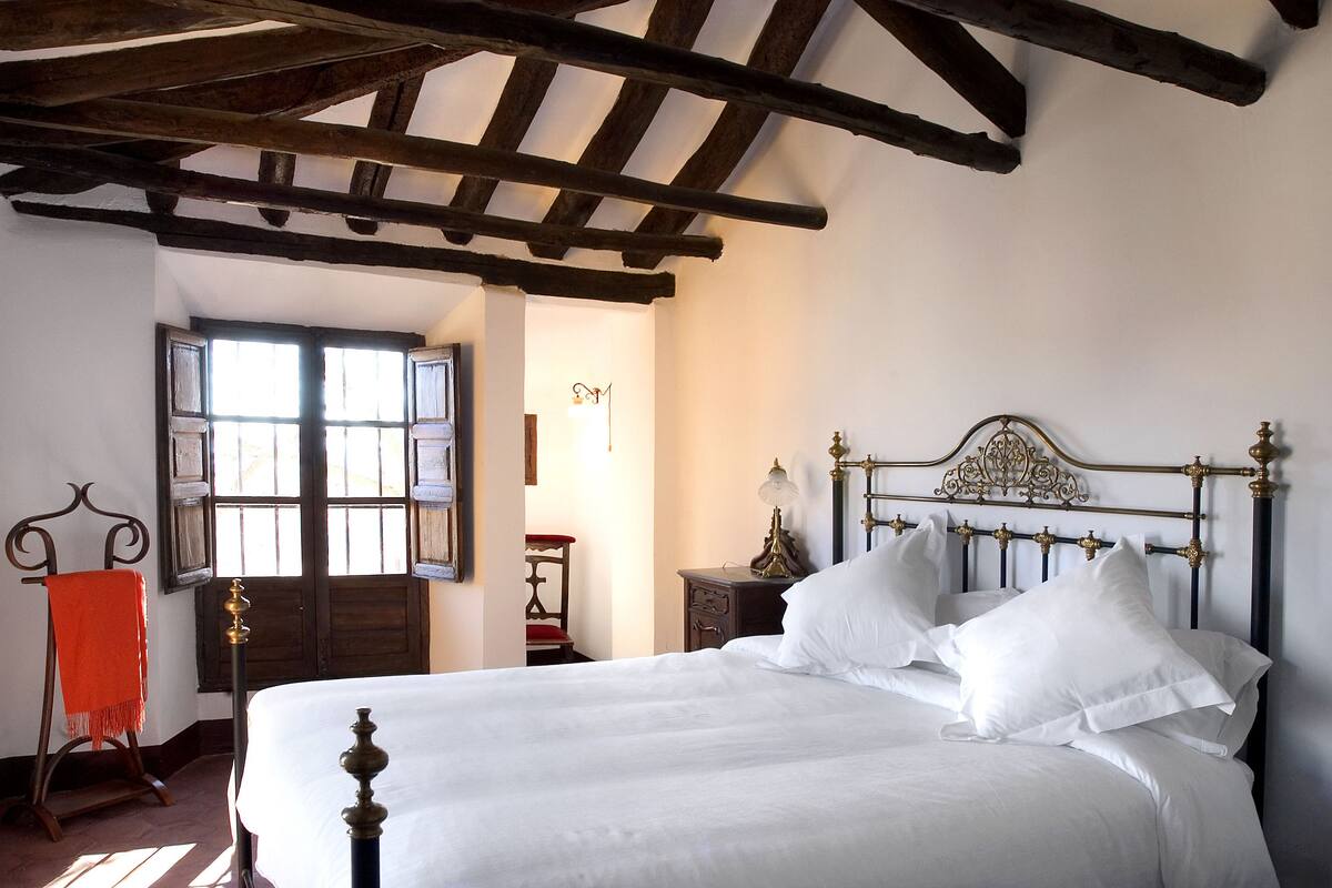 A bedroom with a large bed, wooden ceiling beams, a window with shutters, and a small bedside table with a lamp.