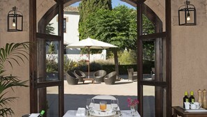 Un comedor con una mesa dispuesta para dos personas, una sombrilla blanca y vistas a un jardín por un ventanal en arco.