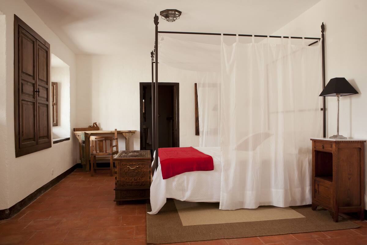 A four-poster bed with a white canopy, a wooden nightstand, a desk, and a chair in a room with white walls and terracotta flooring.