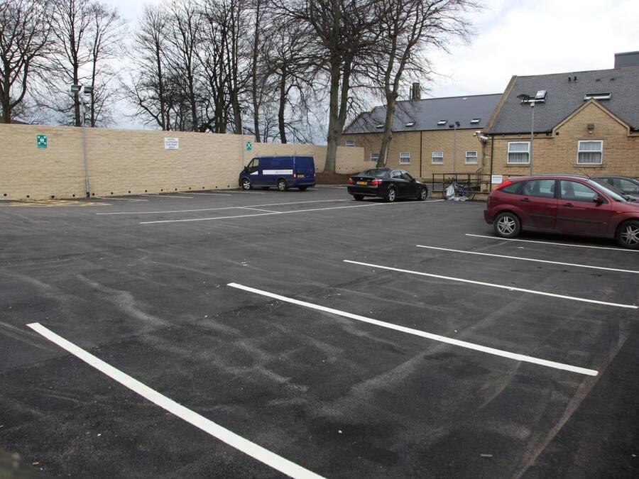 A parking lot with multiple cars and a blue van. There are buildings in the background with a brick wall and a sign indicating a pedestrian path.