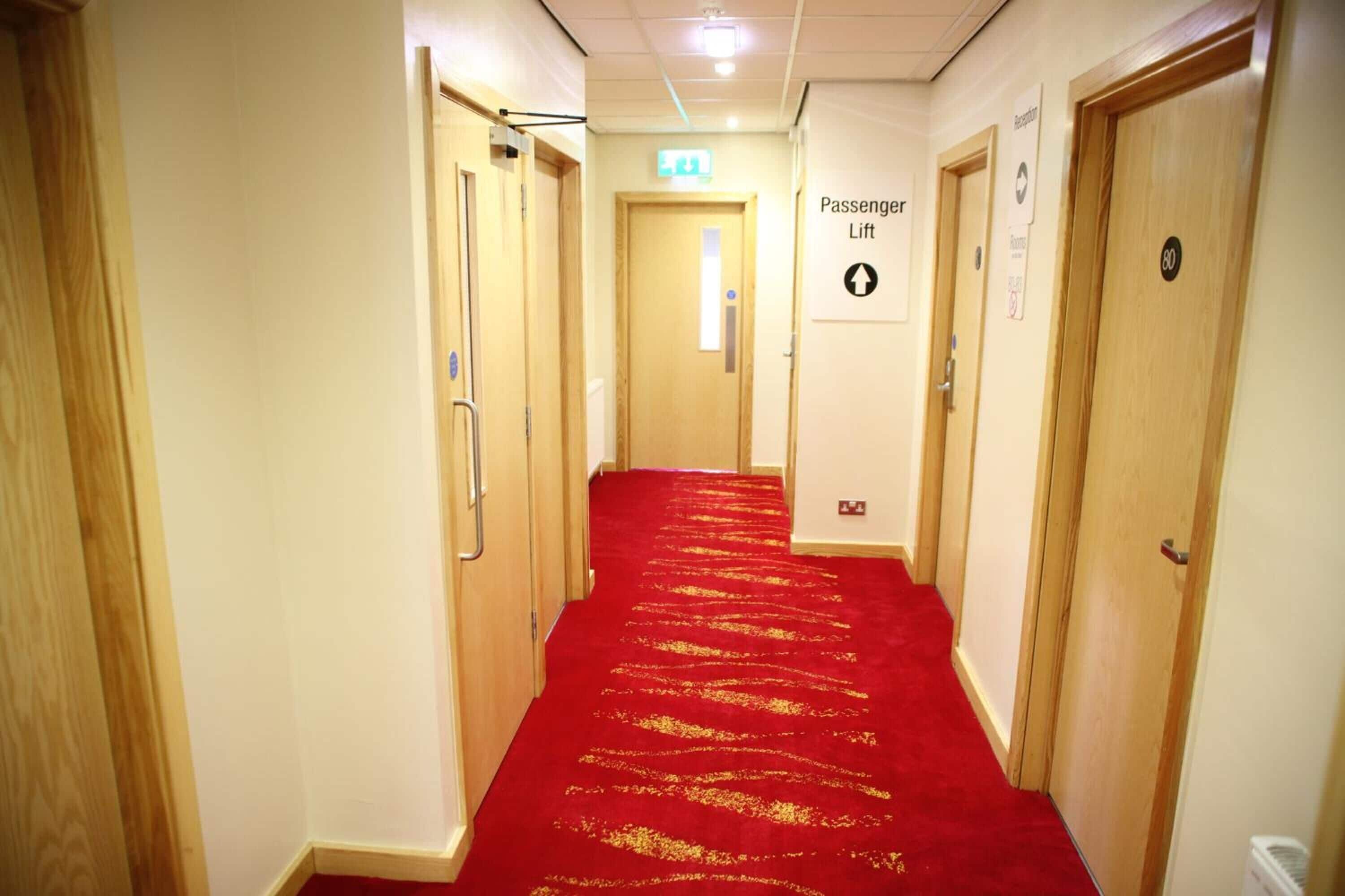 Passadís d'hotel amb una catifa vermella, portes de fusta i un senyal que indica un ascensor.