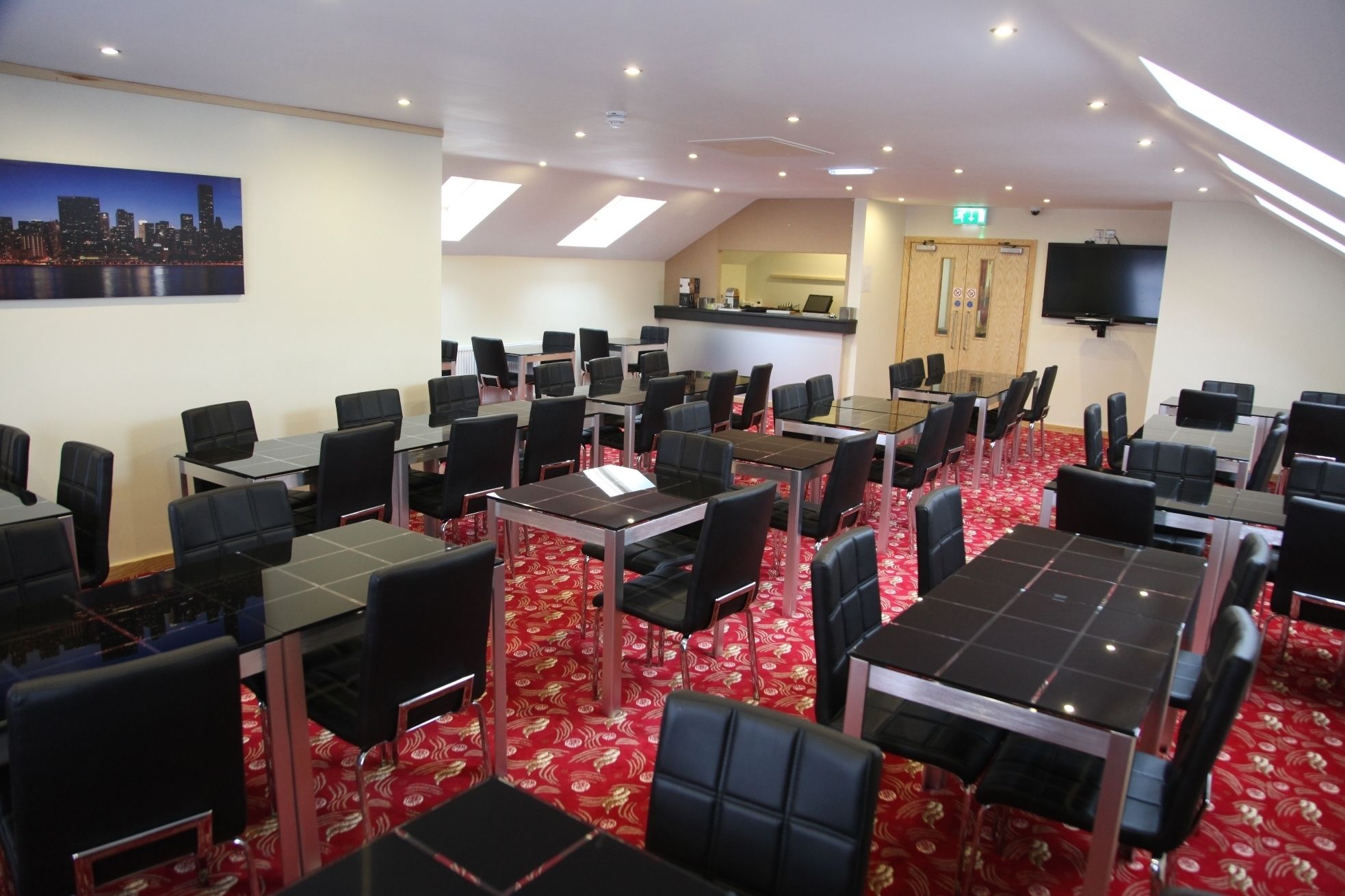 A conference room with multiple tables and chairs, a cityscape painting on the wall, a reception desk, and a television mounted on the wall.